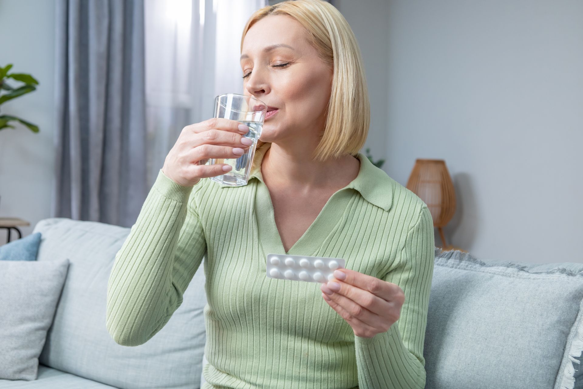 Person in a green sweater holding pills and a glass of water on a couch