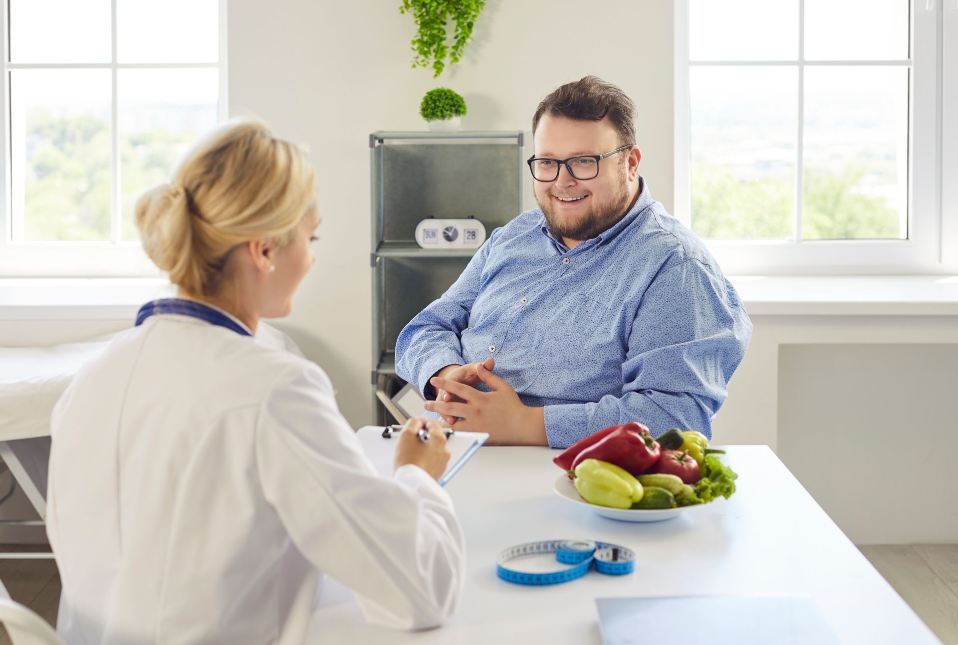 Doctor consulting with seated patient in bright clinic, with fruit and glucose meter on table.