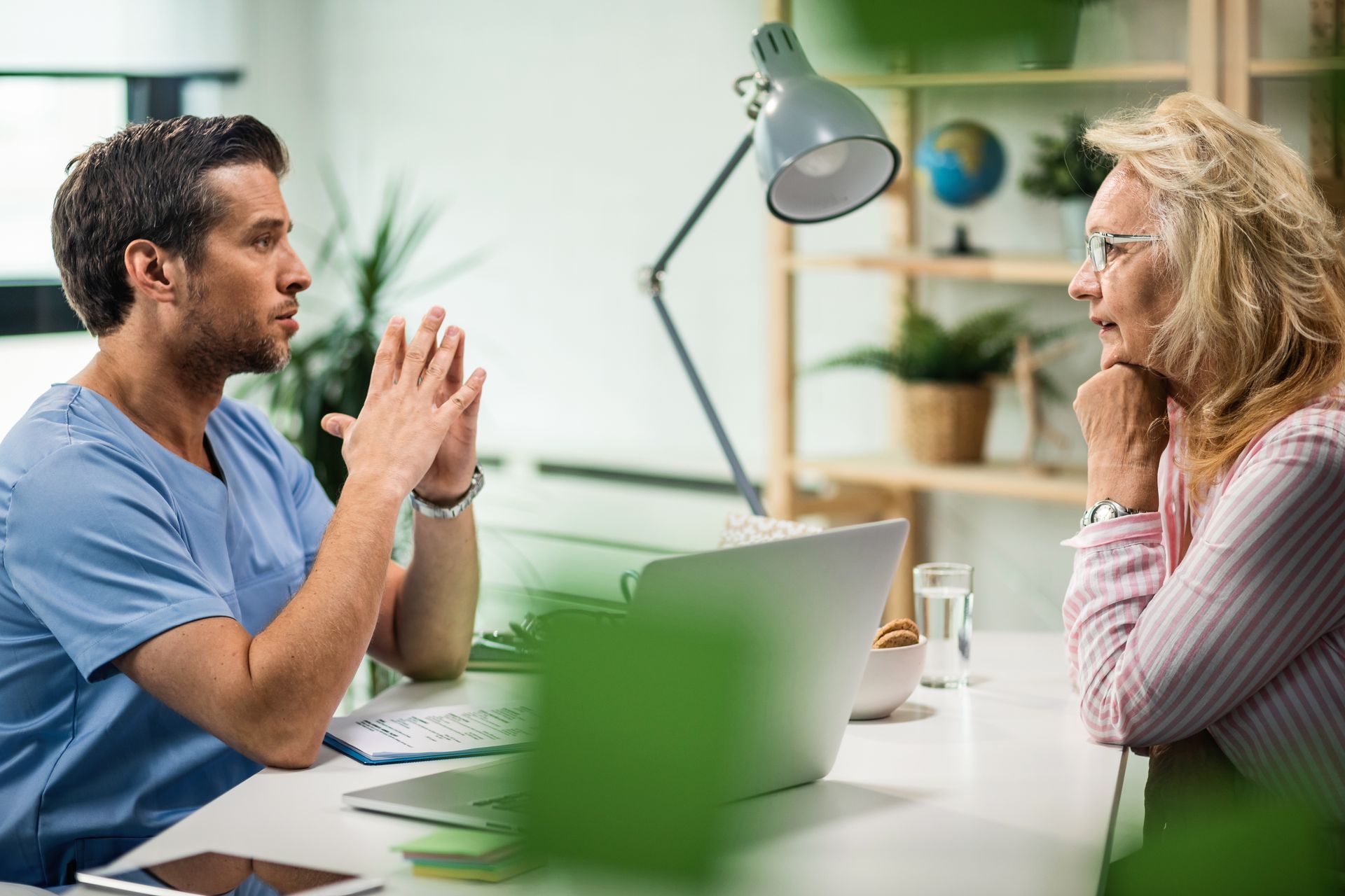 Two people talking across a desk in a bright office, one gesturing while the other listens attentively
