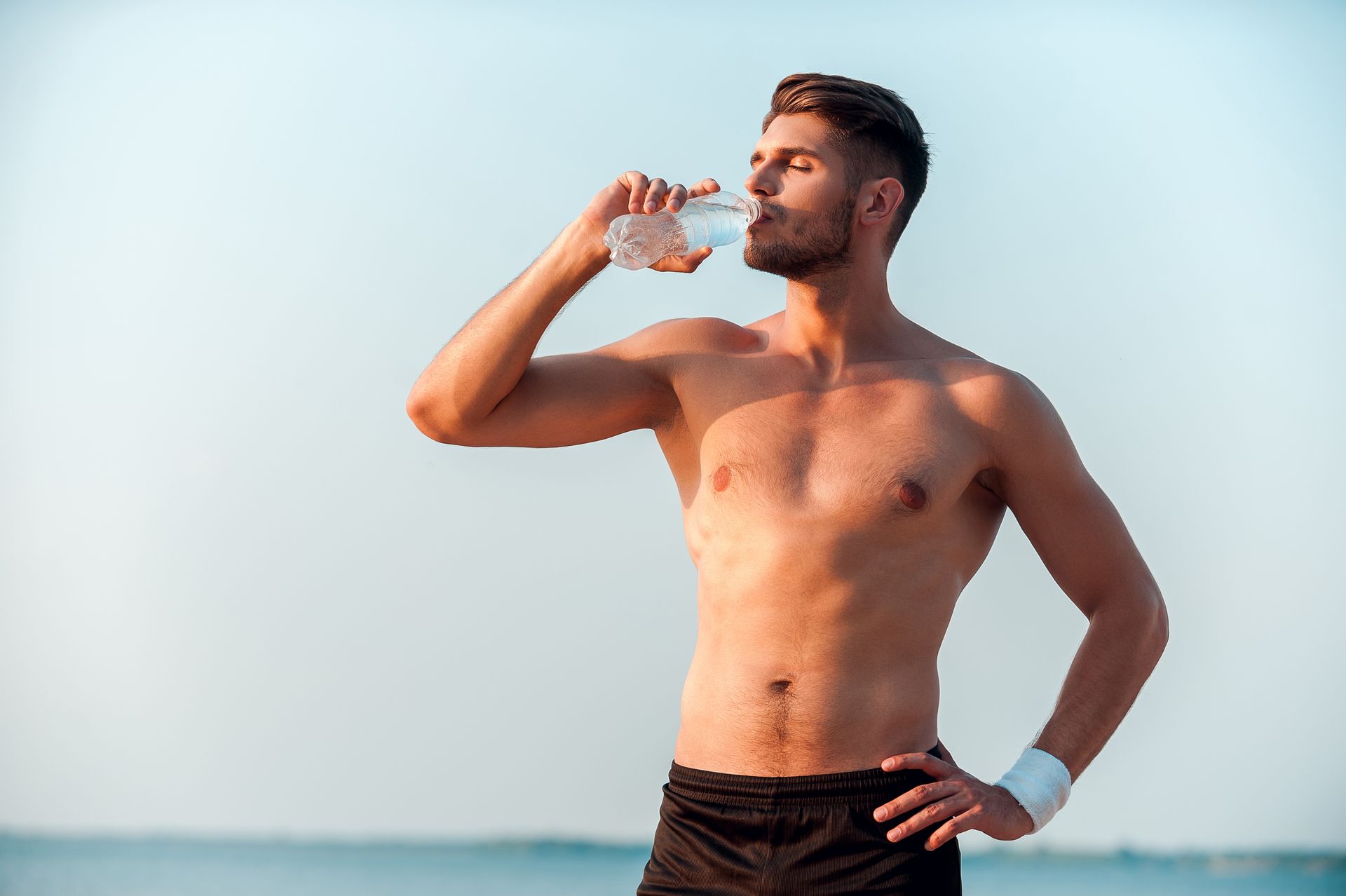 Man standing on a beach drinking water from a bottle in bright sunlight