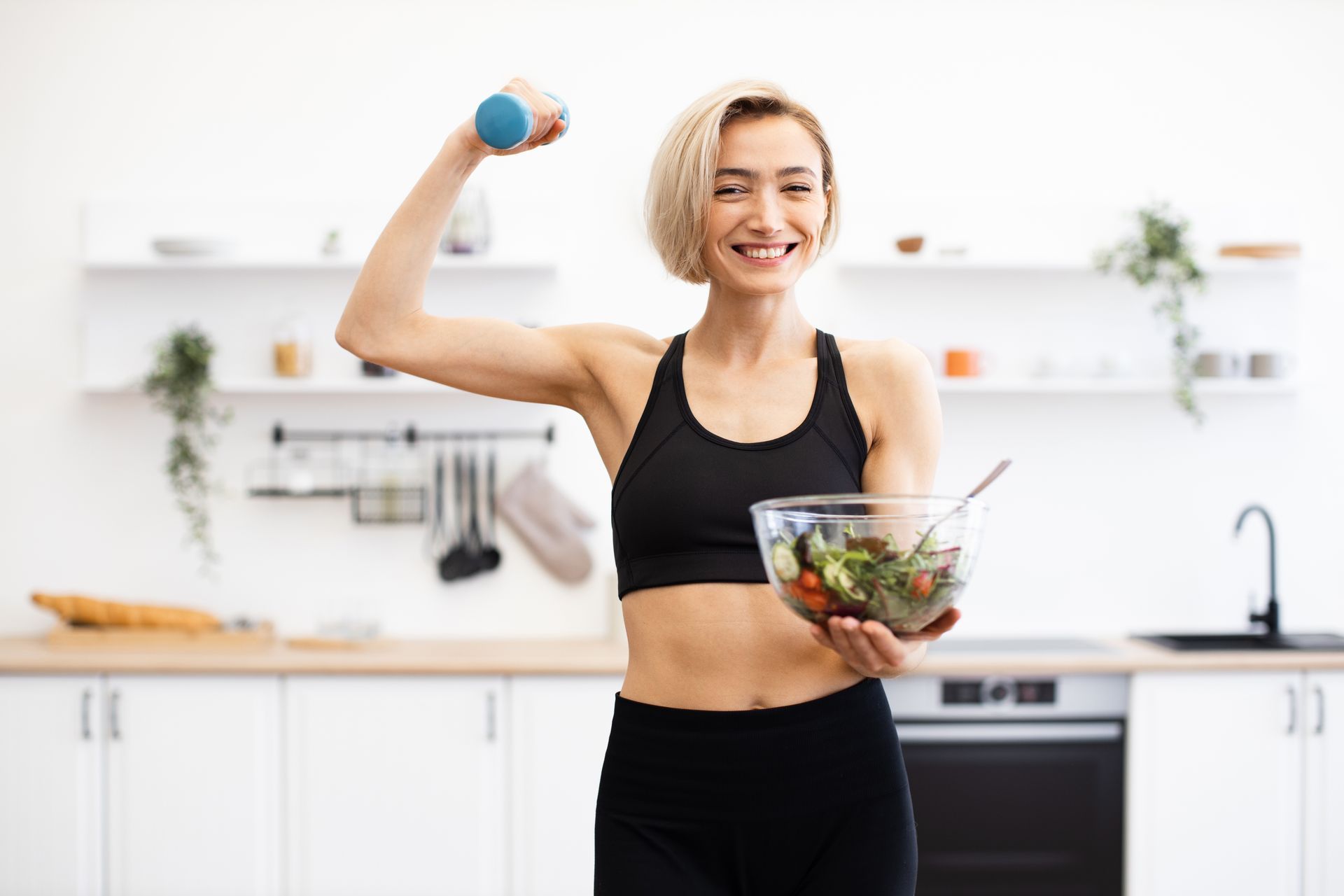 Woman in a bright kitchen holding a salad bowl and a dumbbell, smiling while exercising.
