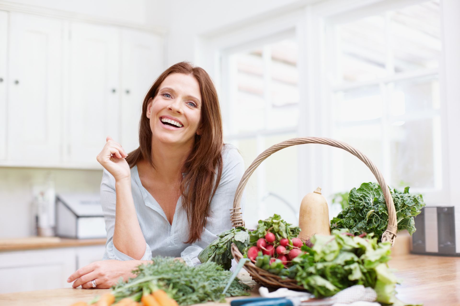 Smiling woman in a bright kitchen beside a basket of fresh vegetables and herbs