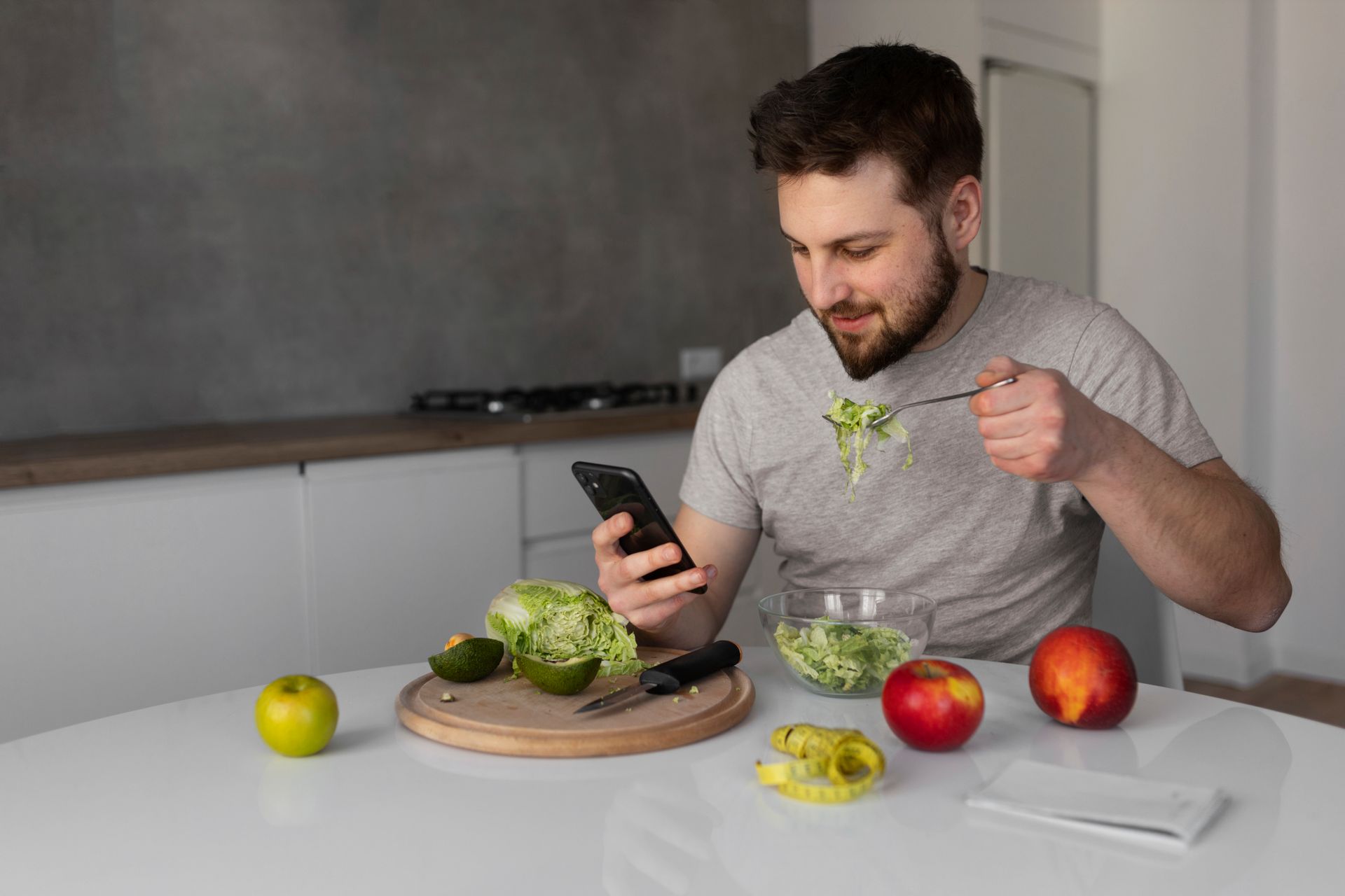 Man in a kitchen photographing a salad while holding a fork, with apples and vegetables on the table