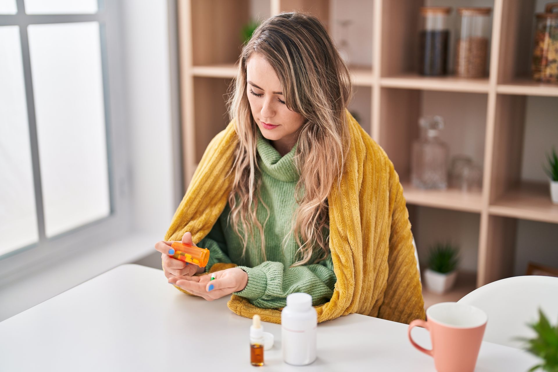 Woman at a table holding pill bottles, with medicine containers and a mug nearby