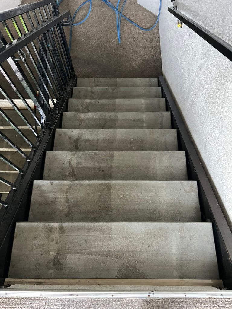Concrete stairs with visible dirt marks are shown. A dark metal railing runs on the left side, and a white wall is on the right, creating an industrial feel.
