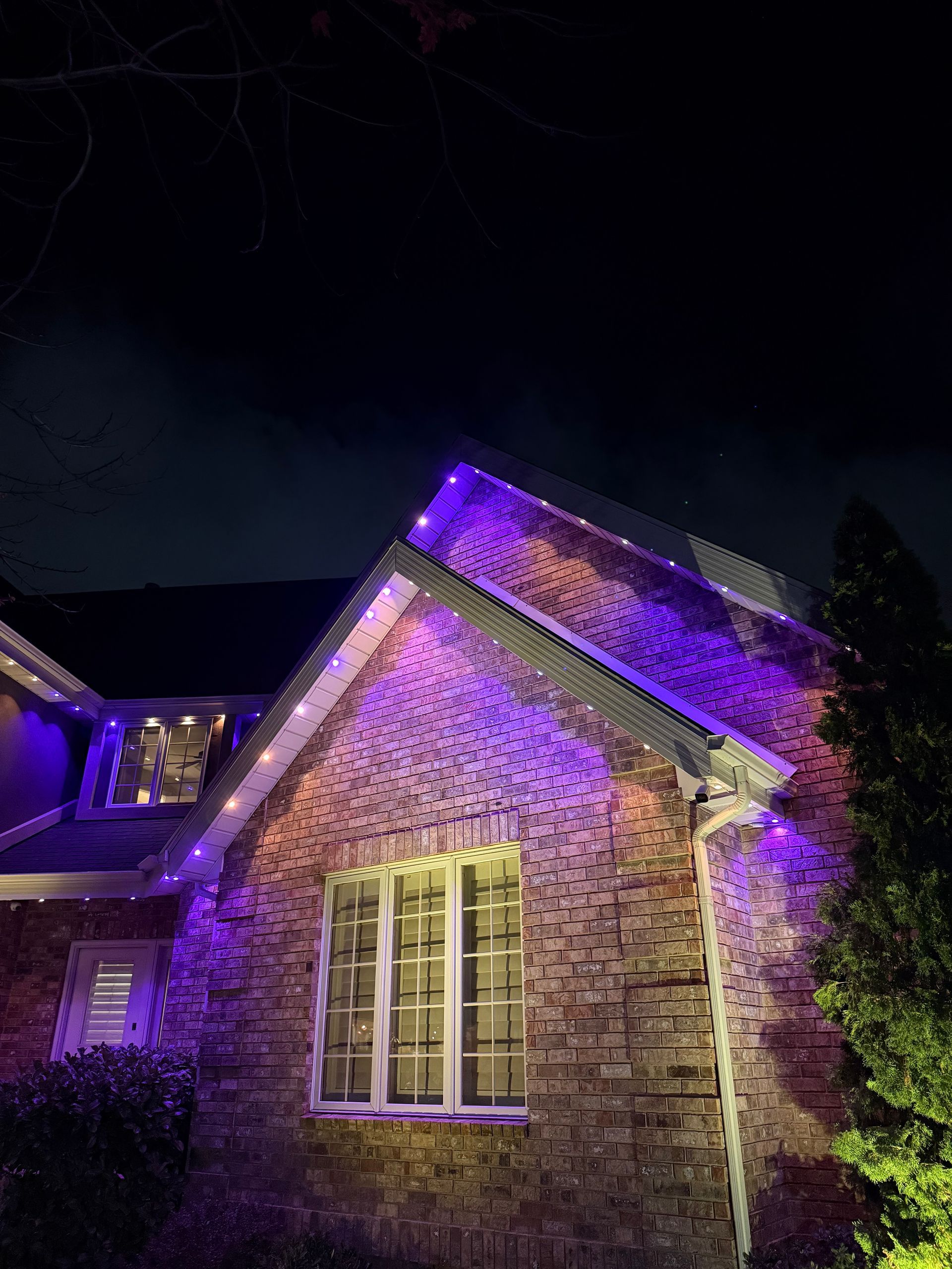 A brick house at night with vibrant purple and white lights outlining the roofline, creating a festive, cozy atmosphere against a dark sky.