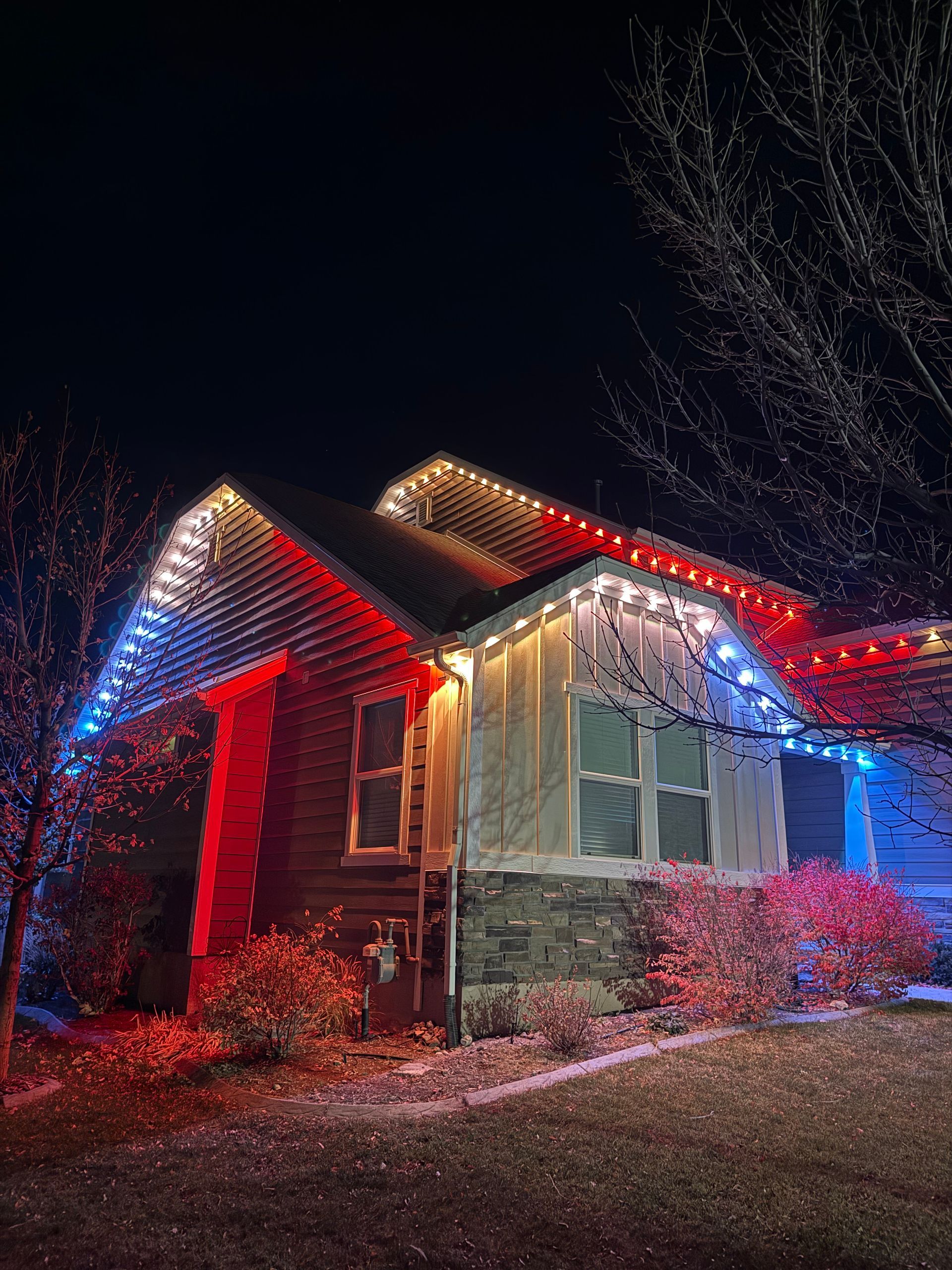 House at night adorned with red, white, and blue lights on the roofline. The vibrant colors illuminate the siding and bare branches, creating a festive atmosphere.