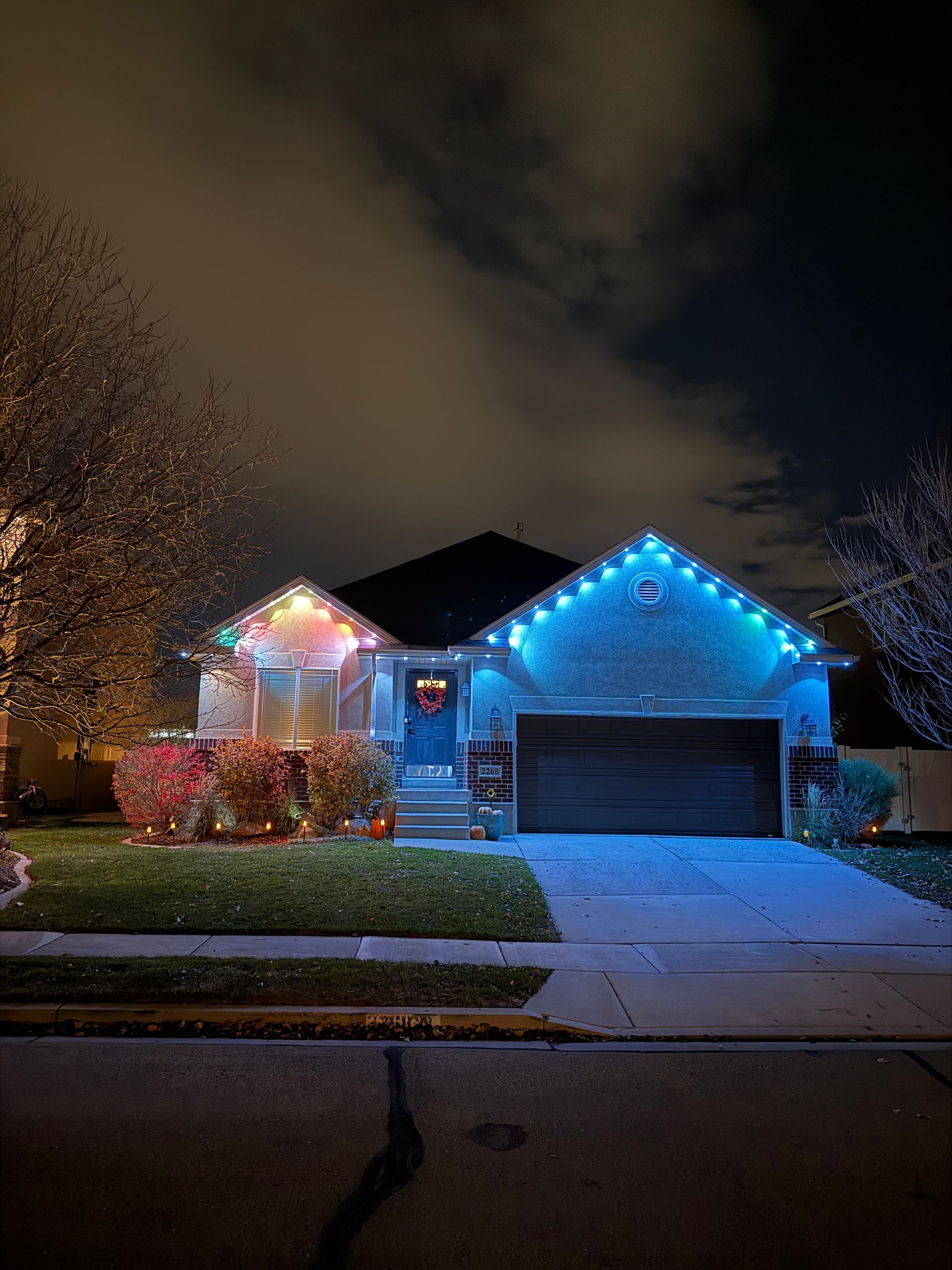 Spacious single-story home at night, adorned with colorful holiday lights on roof and bushes. Warm, festive ambiance with vibrant, cheerful colors.