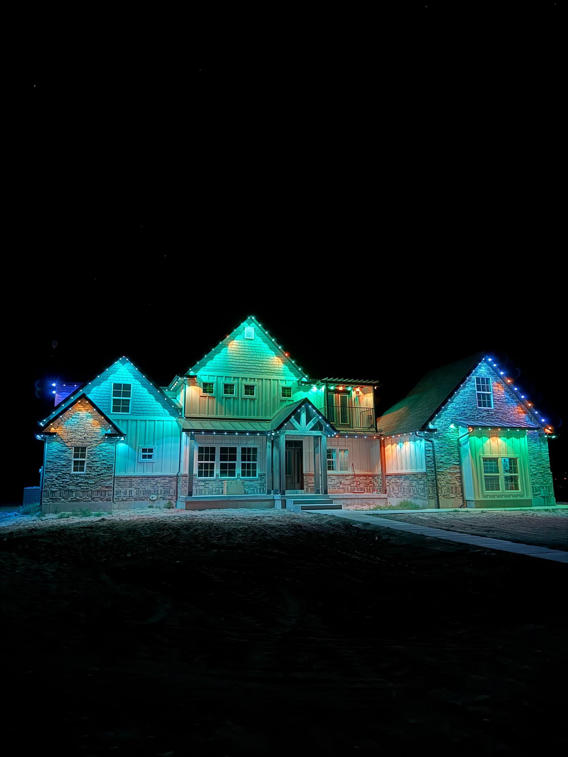 A stone and wood house is adorned with colorful Christmas lights, glowing in green, blue, and red against a dark night sky. The scene feels festive and inviting.
