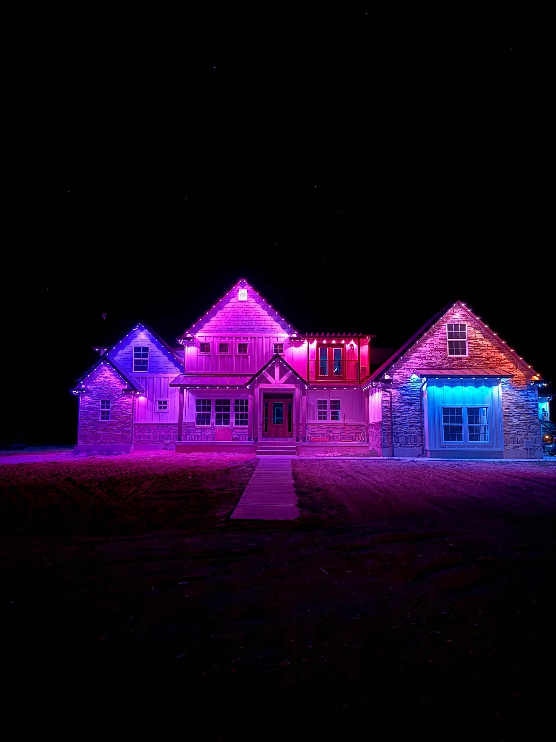A house illuminated at night with vibrant purple, pink, and blue lights, creating a festive and cheerful atmosphere against a dark sky.