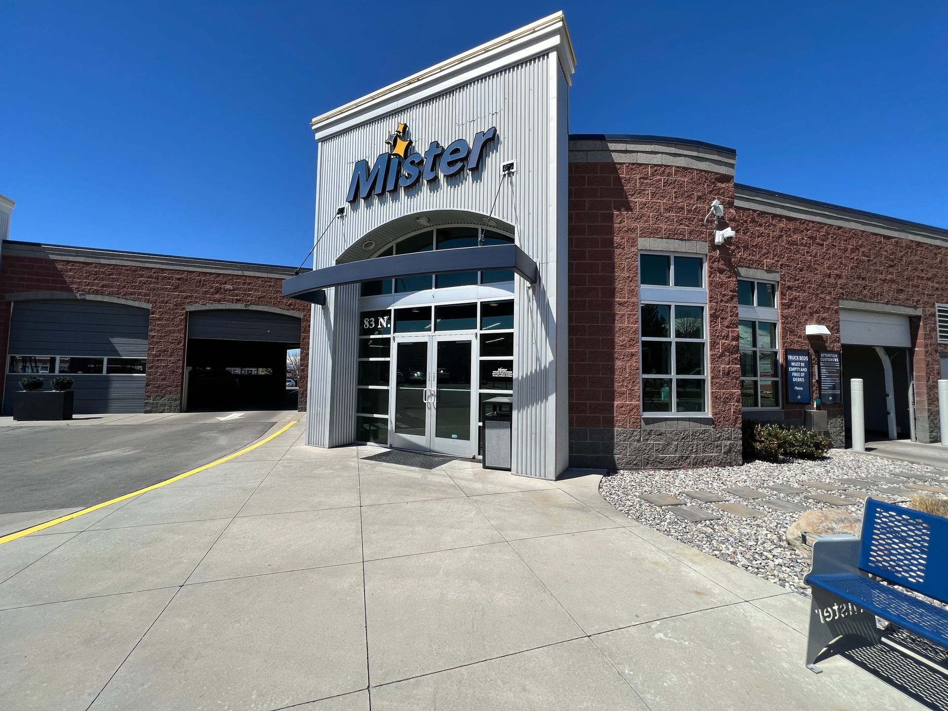 Front view of a Mister Car Wash facility with a modern design: glass entrance, brick walls, and blue sky above. A bench sits to the right.