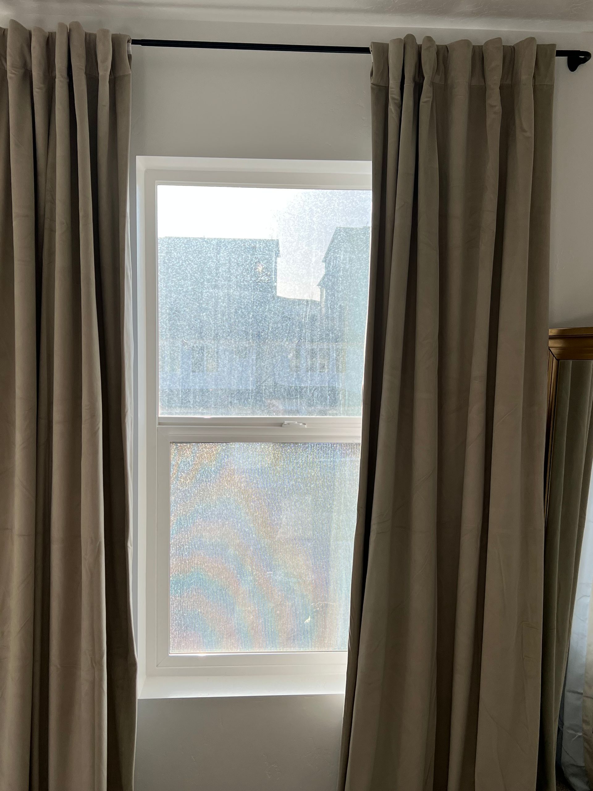A window with taupe curtains open to reveal a rain-splattered glass. Outside, blurred buildings are visible, creating a calm and introspective mood.