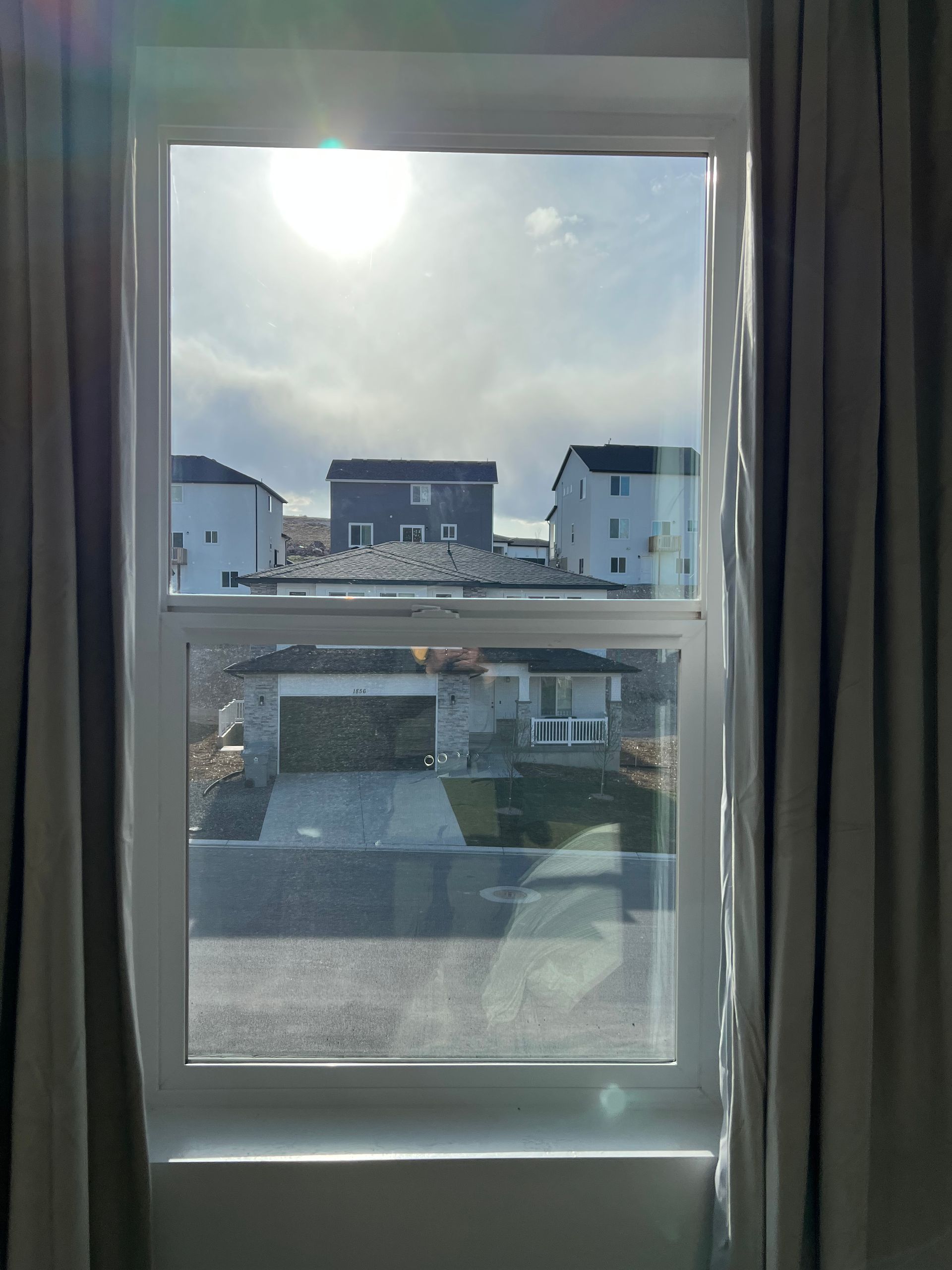 View through a window with gray curtains shows a suburban neighborhood with several modern white and gray houses under a cloudy sky, creating a calm atmosphere.
