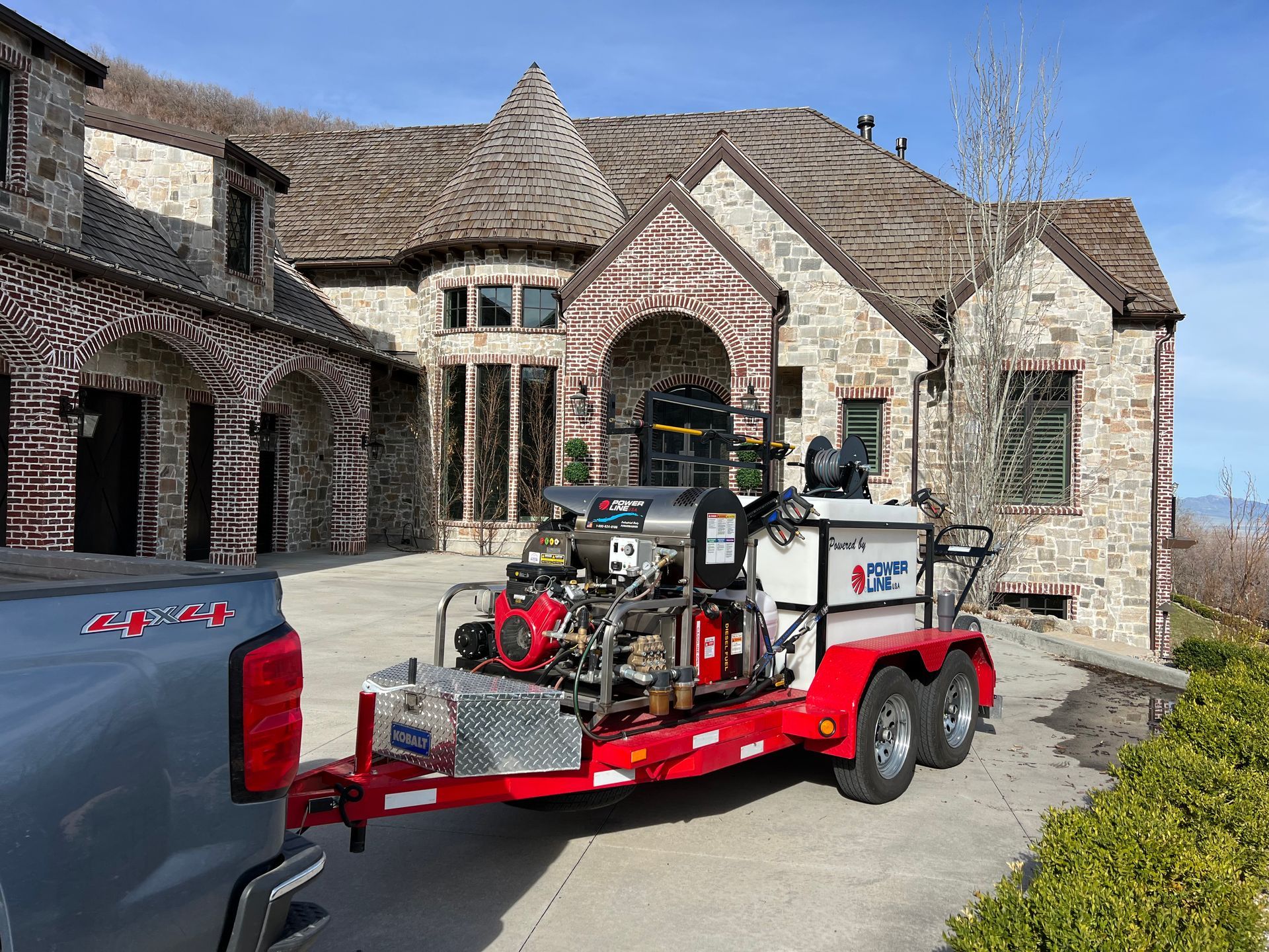 Trailer with industrial equipment hitched to a truck in front of a large stone house. Sunny day, clear sky, with trees and greenery nearby.