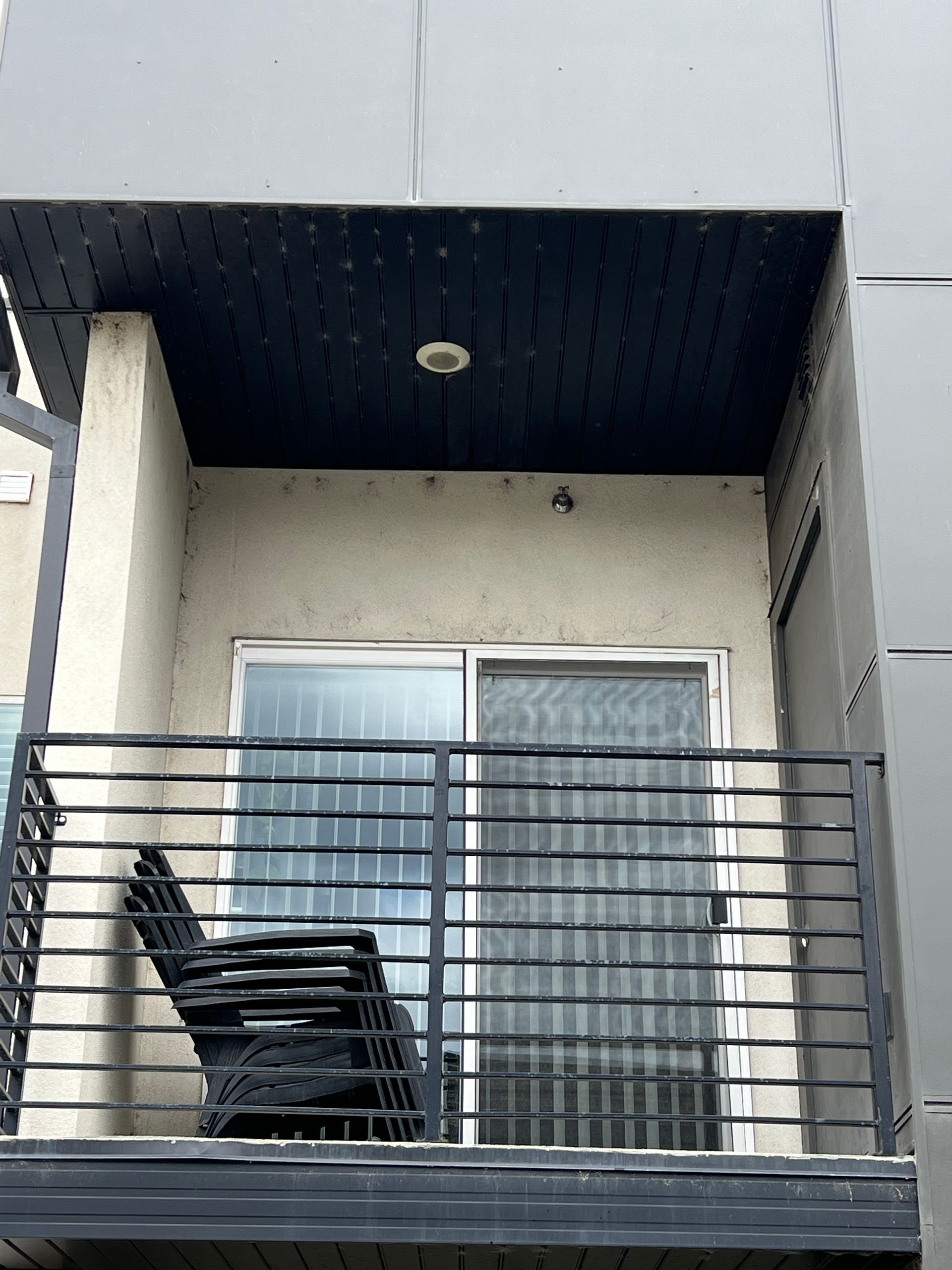 A small balcony with a black metal railing and a single dark chair. Behind, a sliding glass door with vertical blinds partially drawn. The mood is serene.