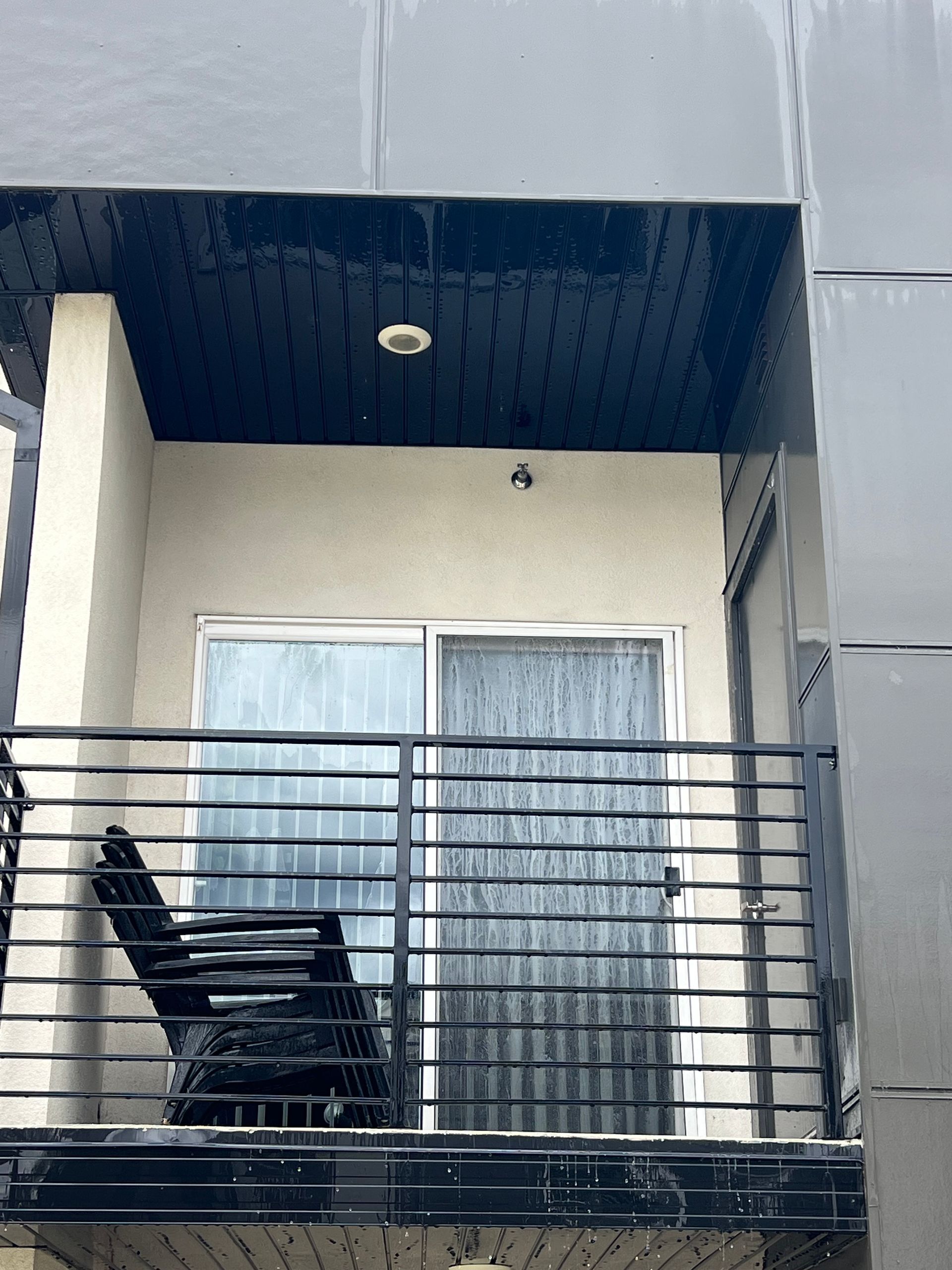 The image shows a small balcony with a metal railing, beige walls, and a black chair. There is a glass sliding door with vertical blinds, conveying a calm urban setting.