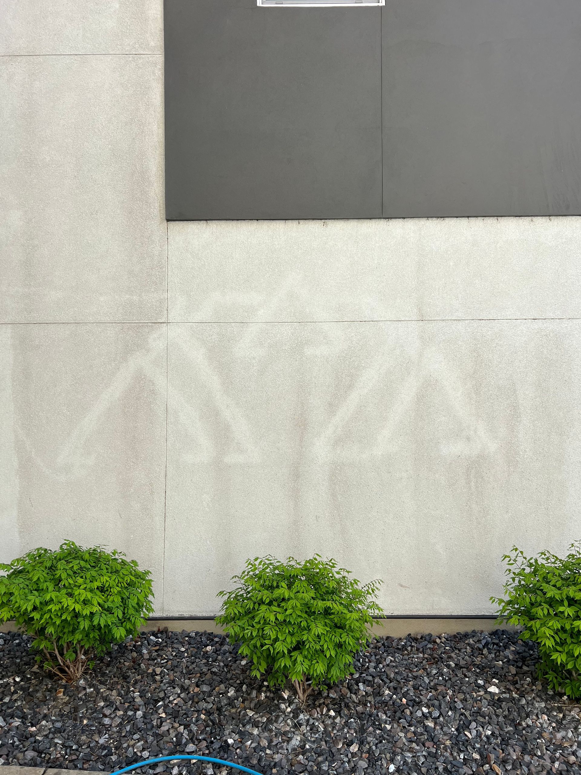 A beige wall with a faint outline of figures, likely caused by cleaning or weathering, above a row of green shrubs. The tone is calm and understated.