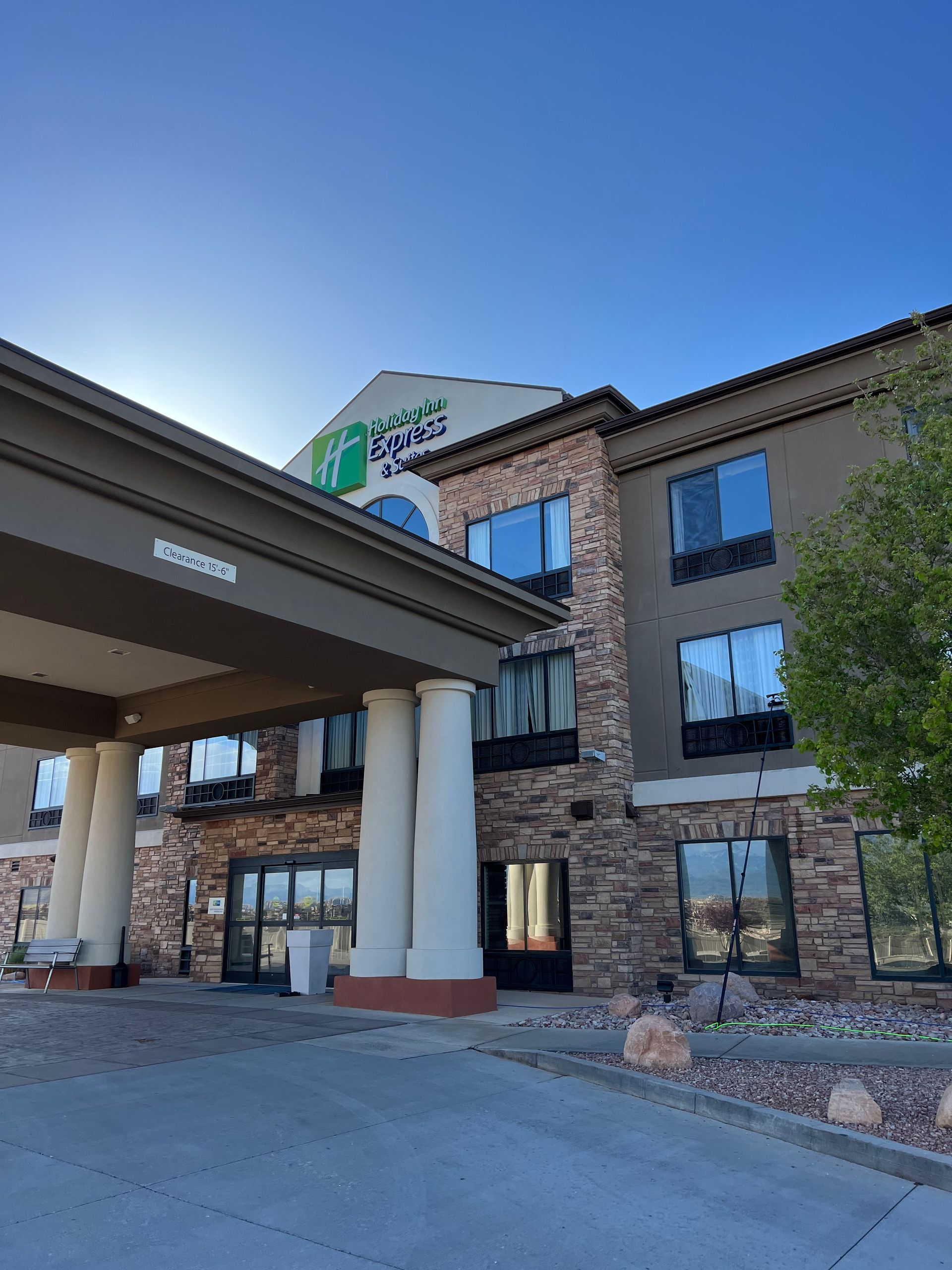 The image shows the exterior of a three-story hotel with brick and stucco facade under a clear blue sky. A tree partially frames the building.