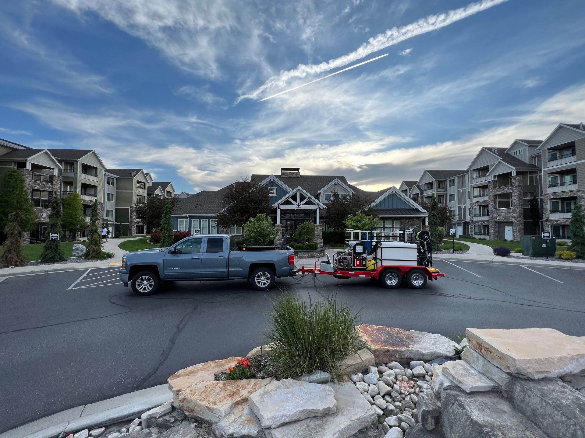 A gray pickup truck with a red trailer is parked in front of a modern apartment complex under a dynamic sky. The scene feels calm and orderly.