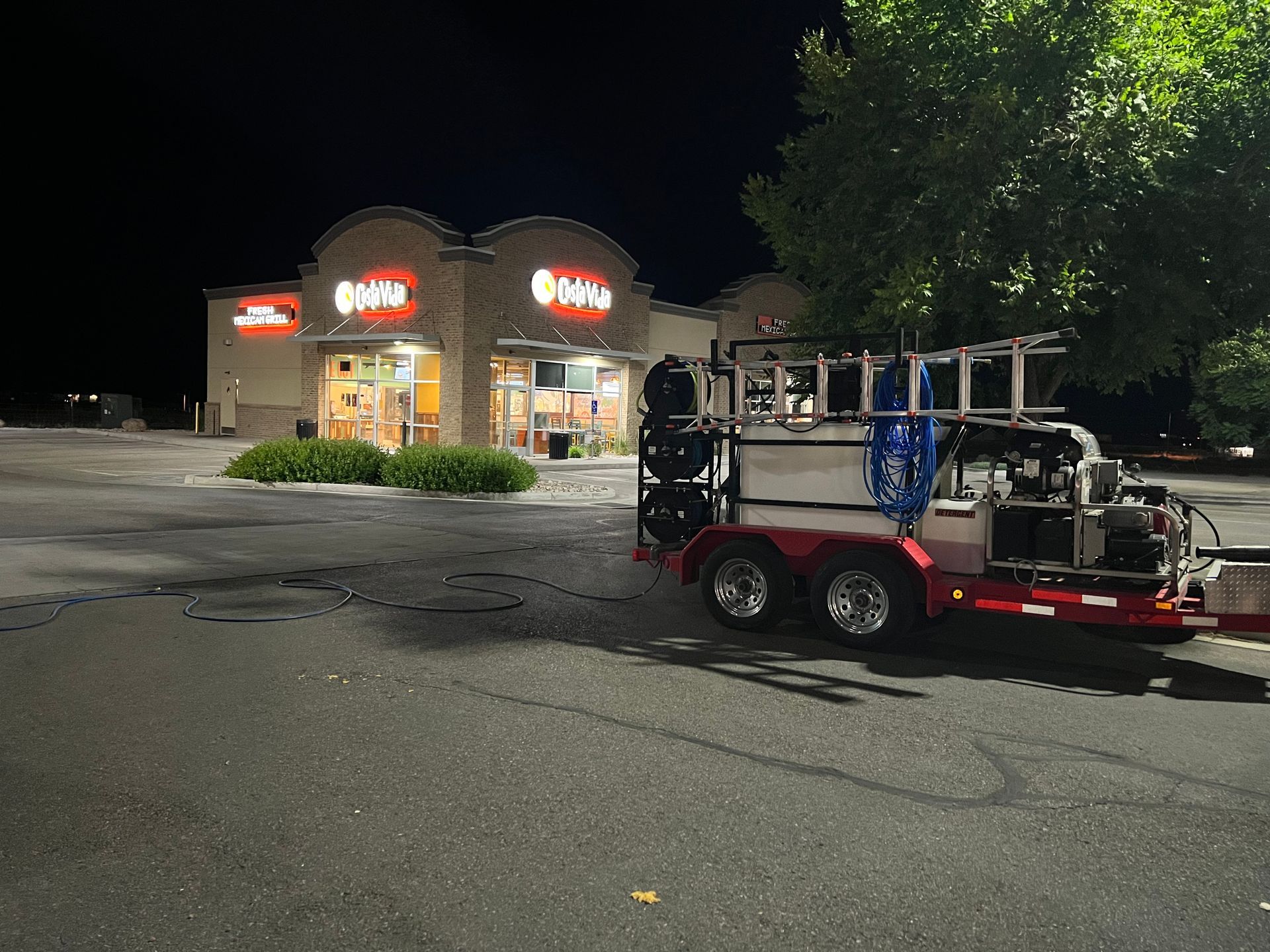 A well-lit convenience store is seen at night, with bright signs and shrubbery in front. A red utility trailer with equipment is parked under a tree nearby.