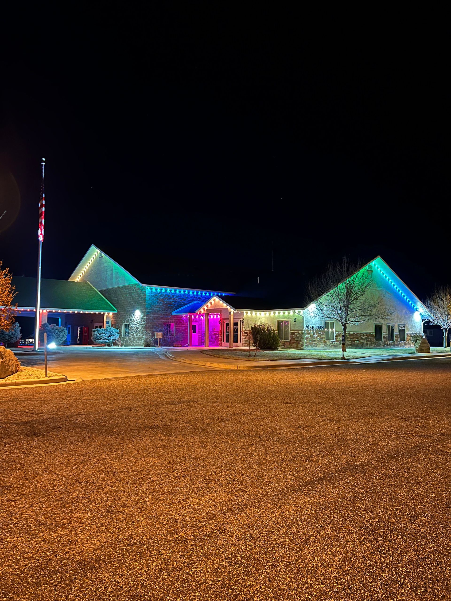 A building at night adorned with multicolored Christmas lights, including green, blue, and pink. A flagpole stands in the foreground, adding a festive ambiance.