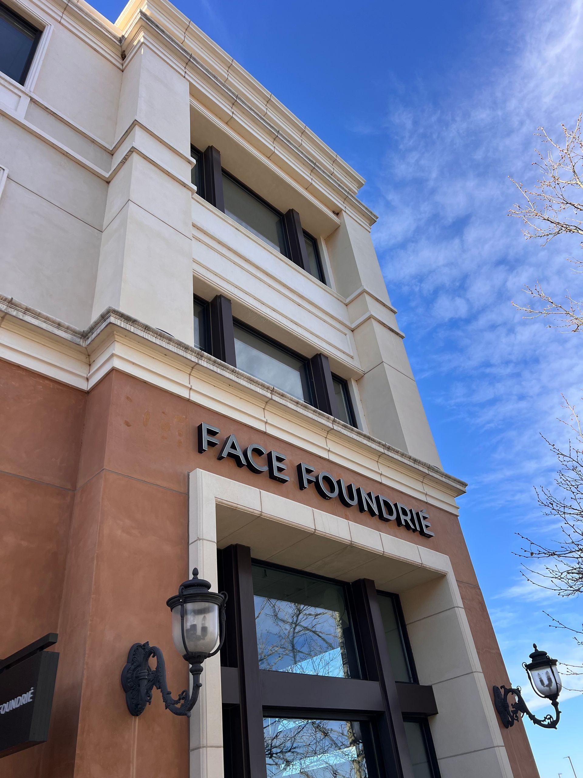 A building facade with a partially painted brown wall and exposed beige sections. A ladder leans against it, near large windows and a sign reading