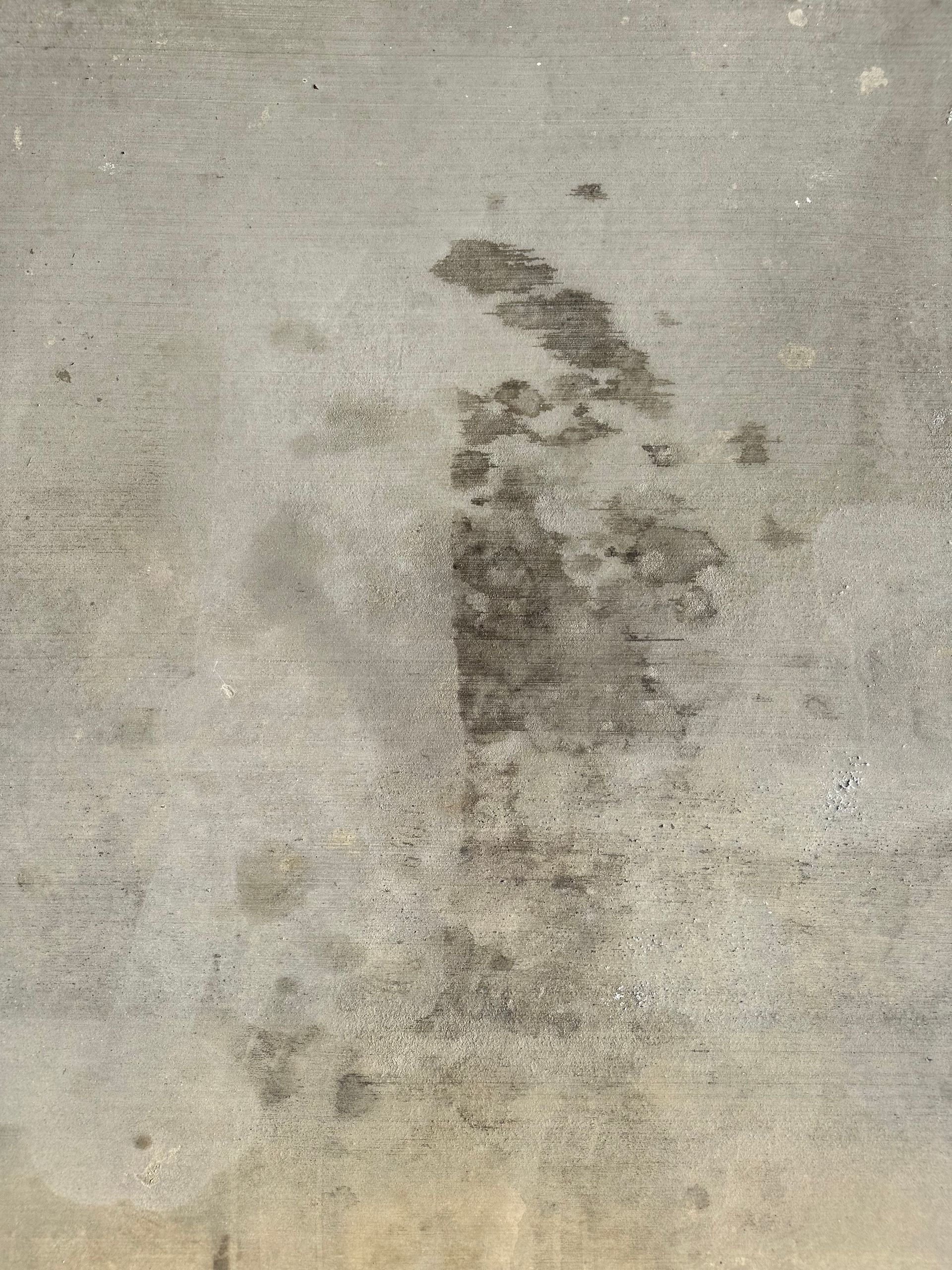 Rough concrete surface with dark stains creating abstract patterns. The gray backdrop and irregular textures convey an industrial, gritty feel.