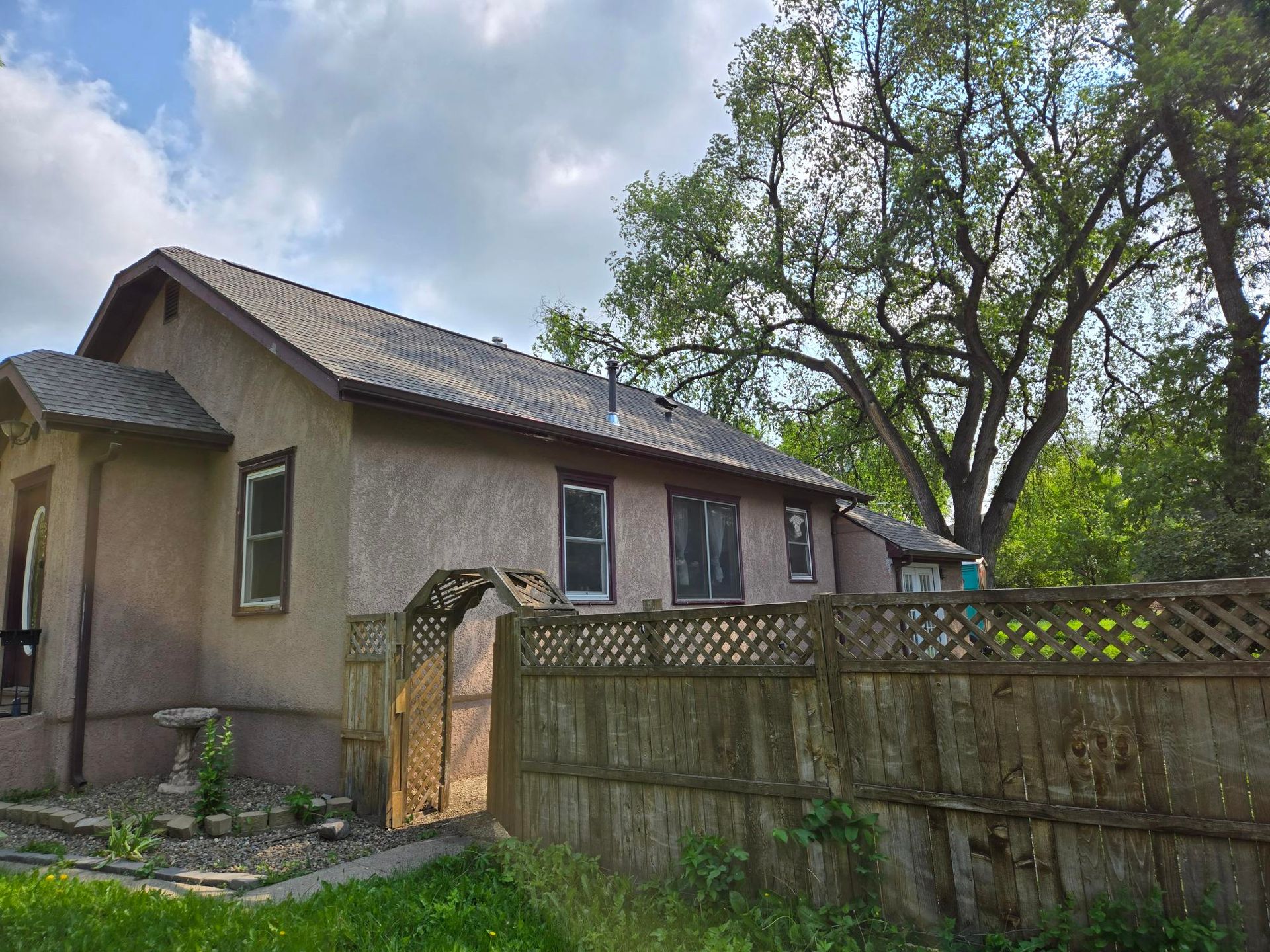 Beige house with a sloped roof, behind a wooden fence and archway, with a large tree in the background.