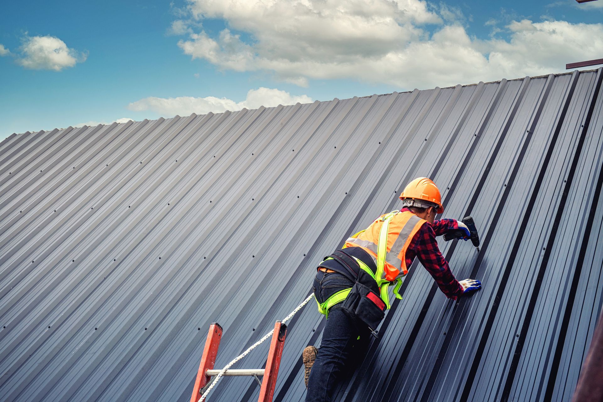 Roofer in safety gear installing metal panels on a roof against a cloudy sky.