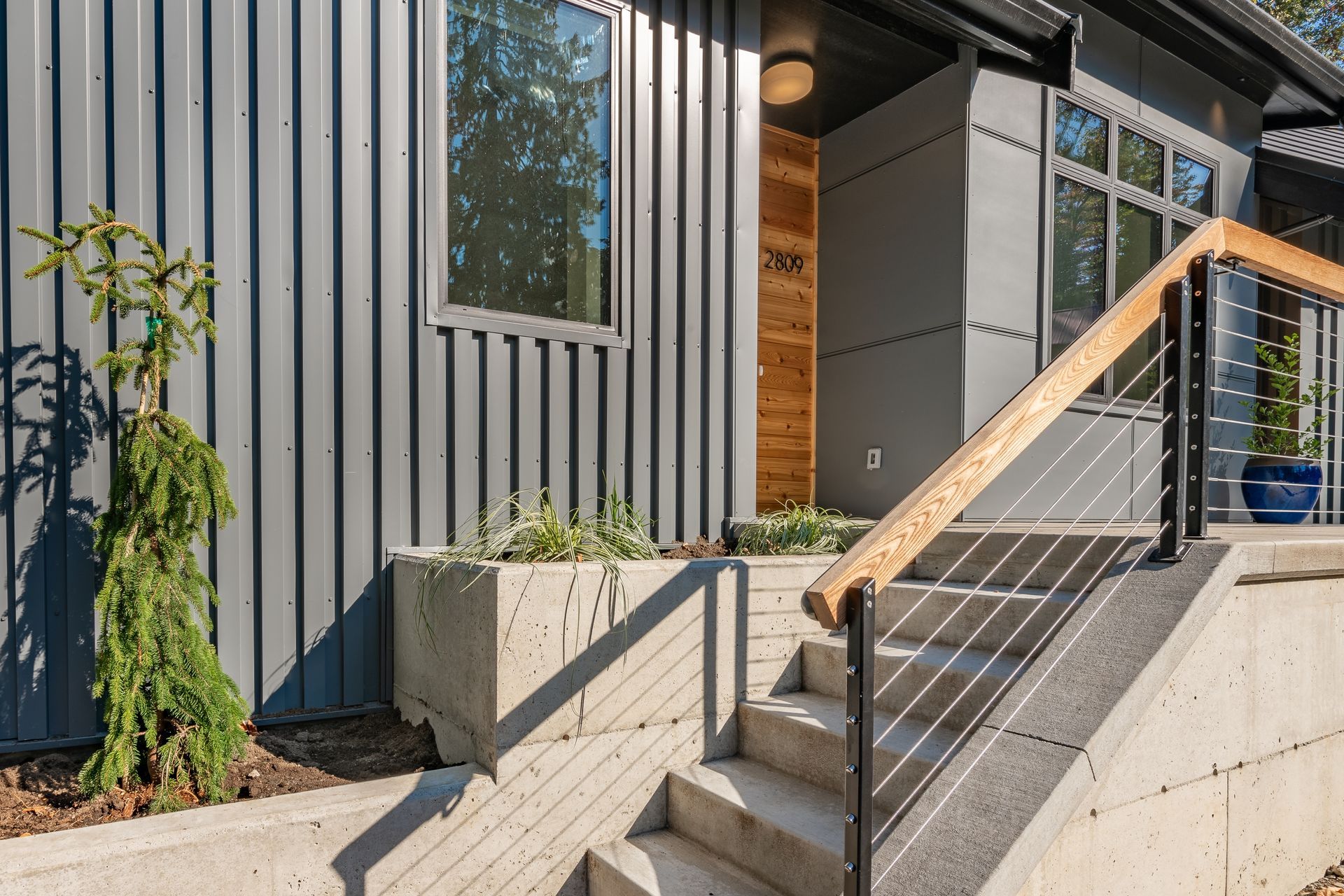 Modern home exterior with gray siding, concrete stairs, wooden door, and steel cable railing.