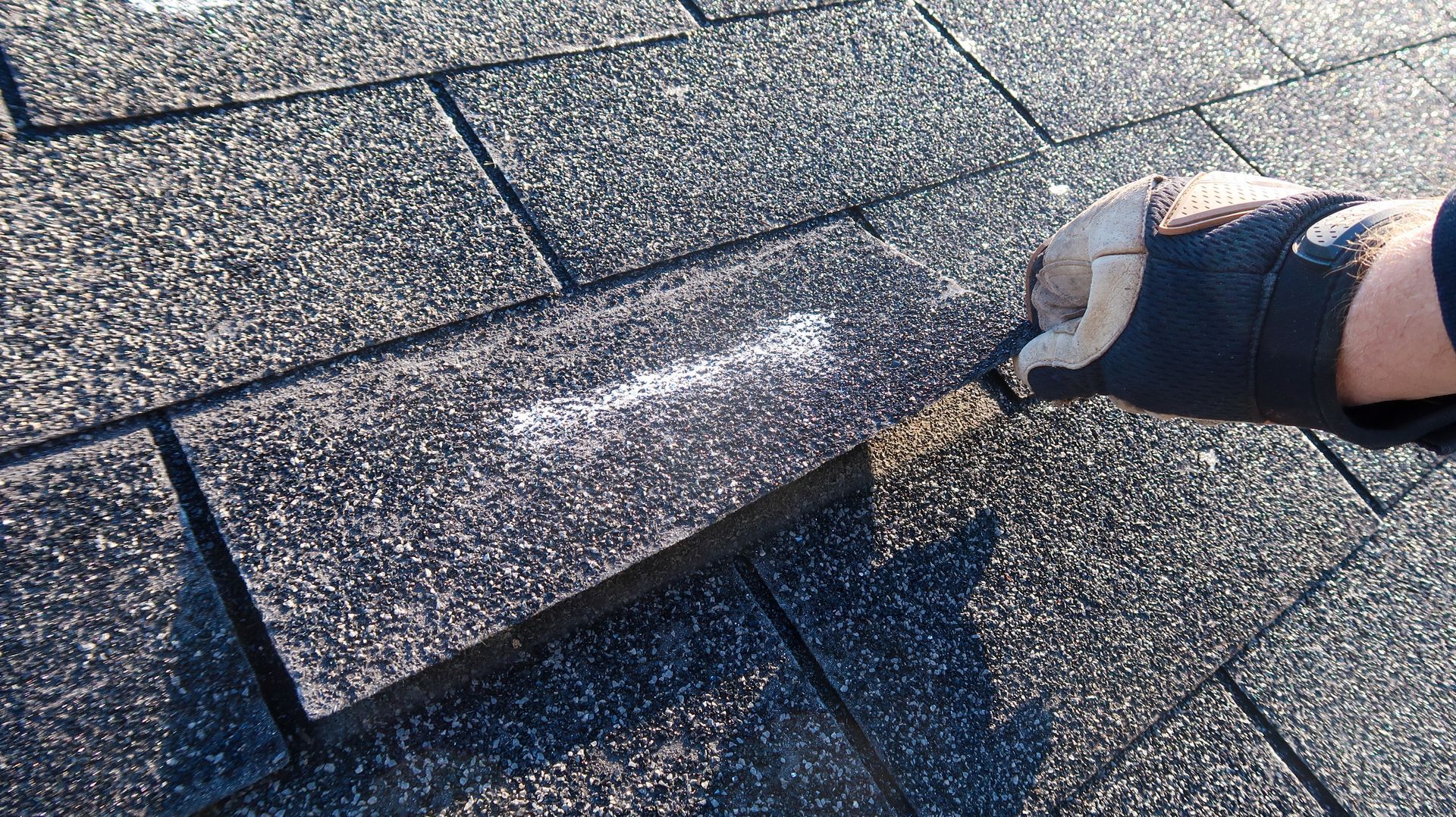 Gloved hand holds asphalt roof shingle with white marking, set on other shingles.