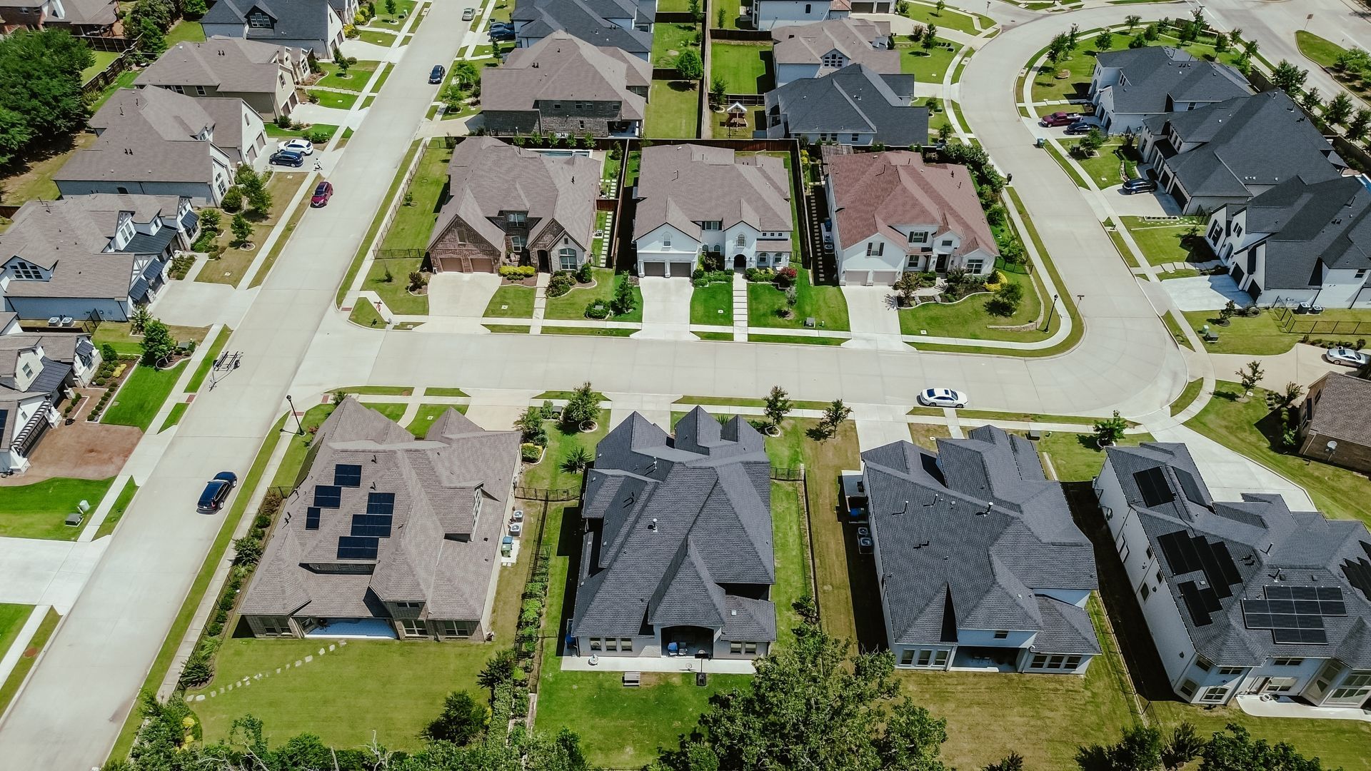 Aerial view of residential neighborhood with houses, streets, and green lawns under a sunny sky.