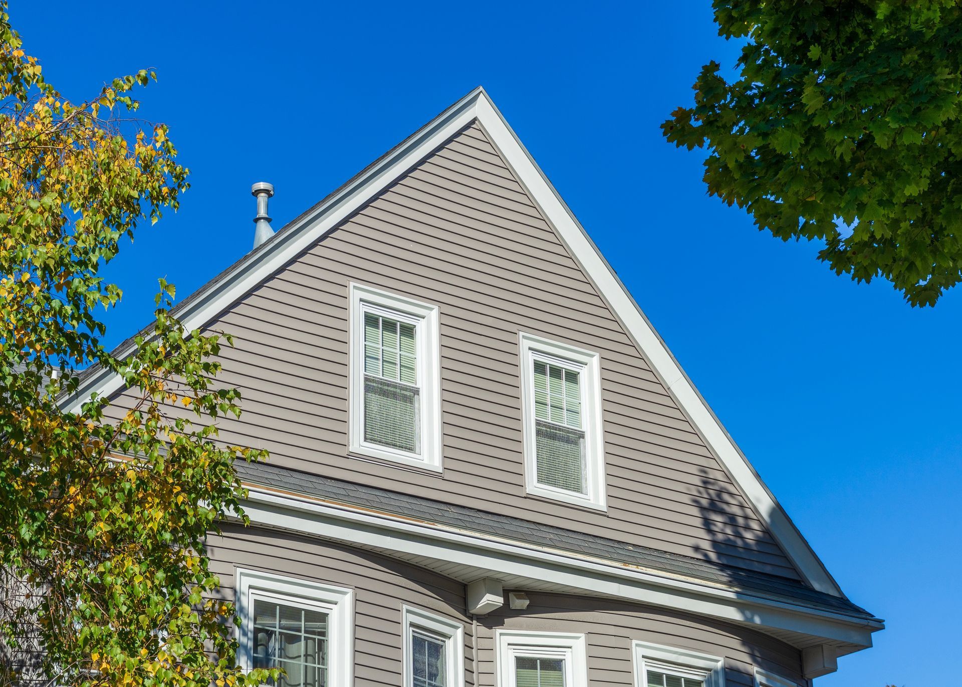 Gray house with white-framed windows, against a bright blue sky, with autumn trees on either side.