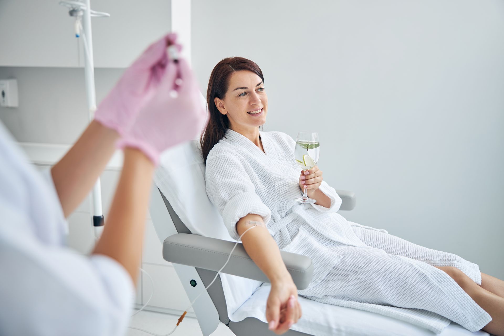 Woman in robe gets IV infusion while holding drink, smiling in a light-filled medical setting.