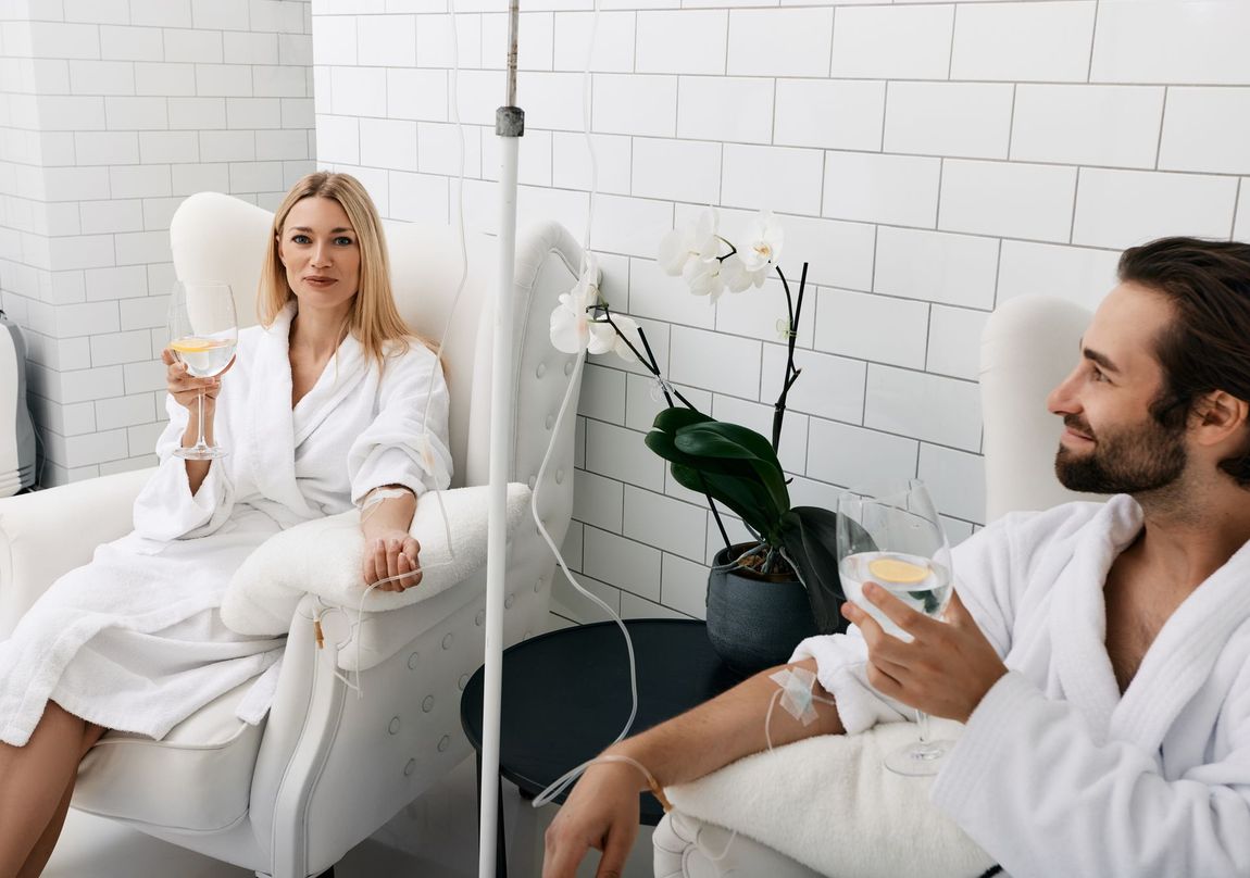 A man and a woman are sitting in chairs with iv drips in their hands.