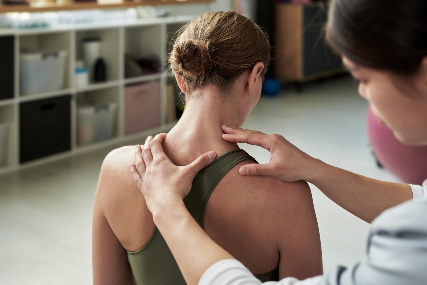 Person's shoulder and neck being examined by another person's hands. Indoor setting, medical examination.