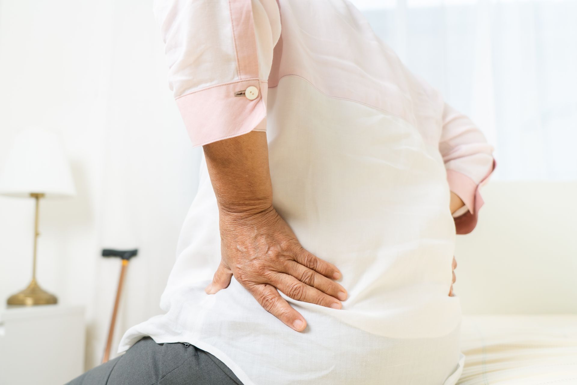 An elderly woman is sitting on a bed holding her back in pain.