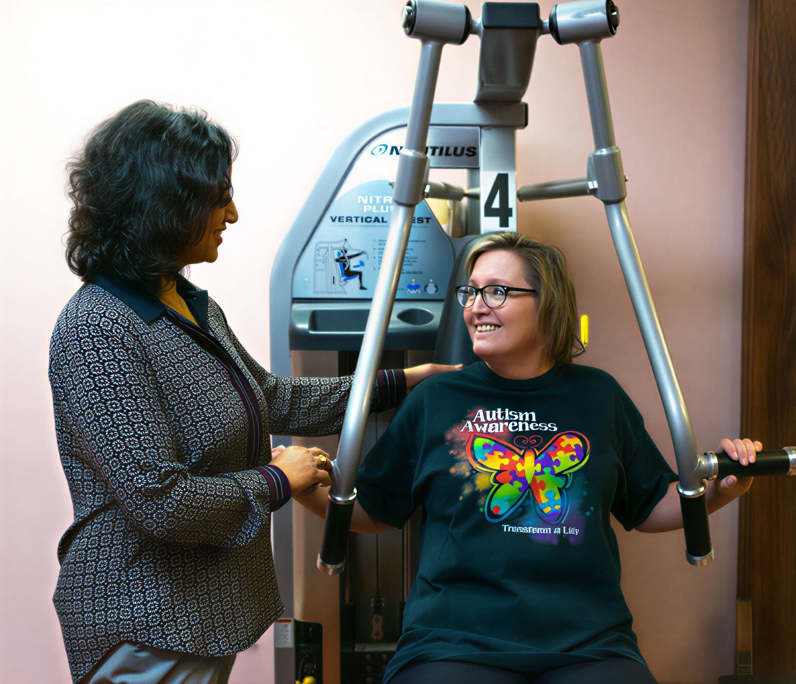 Woman using chest press machine, assisted by another woman. They are in a gym.