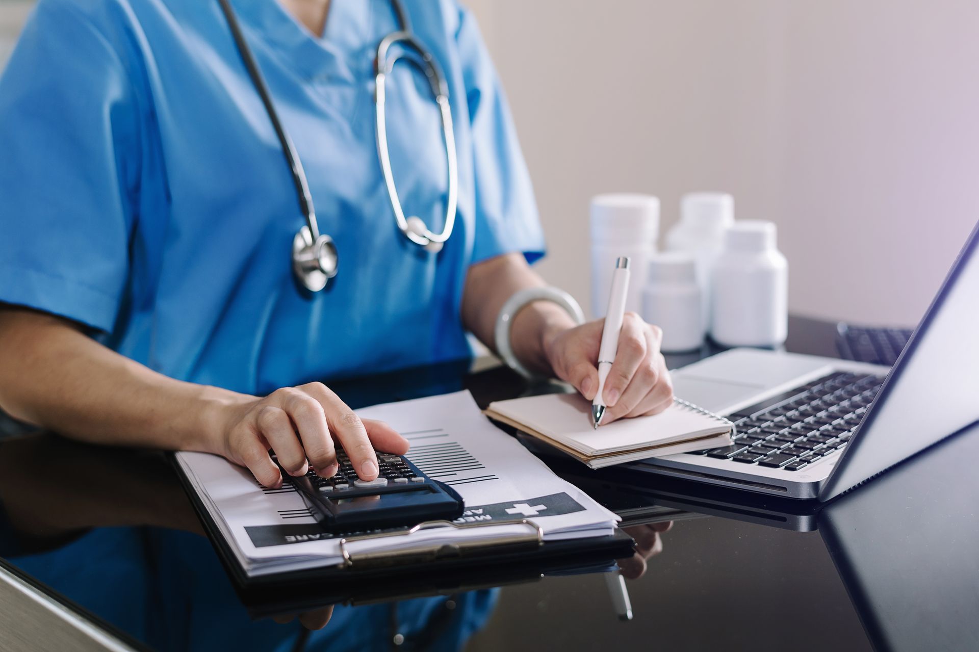 A nurse is sitting at a desk using a calculator and a laptop computer.