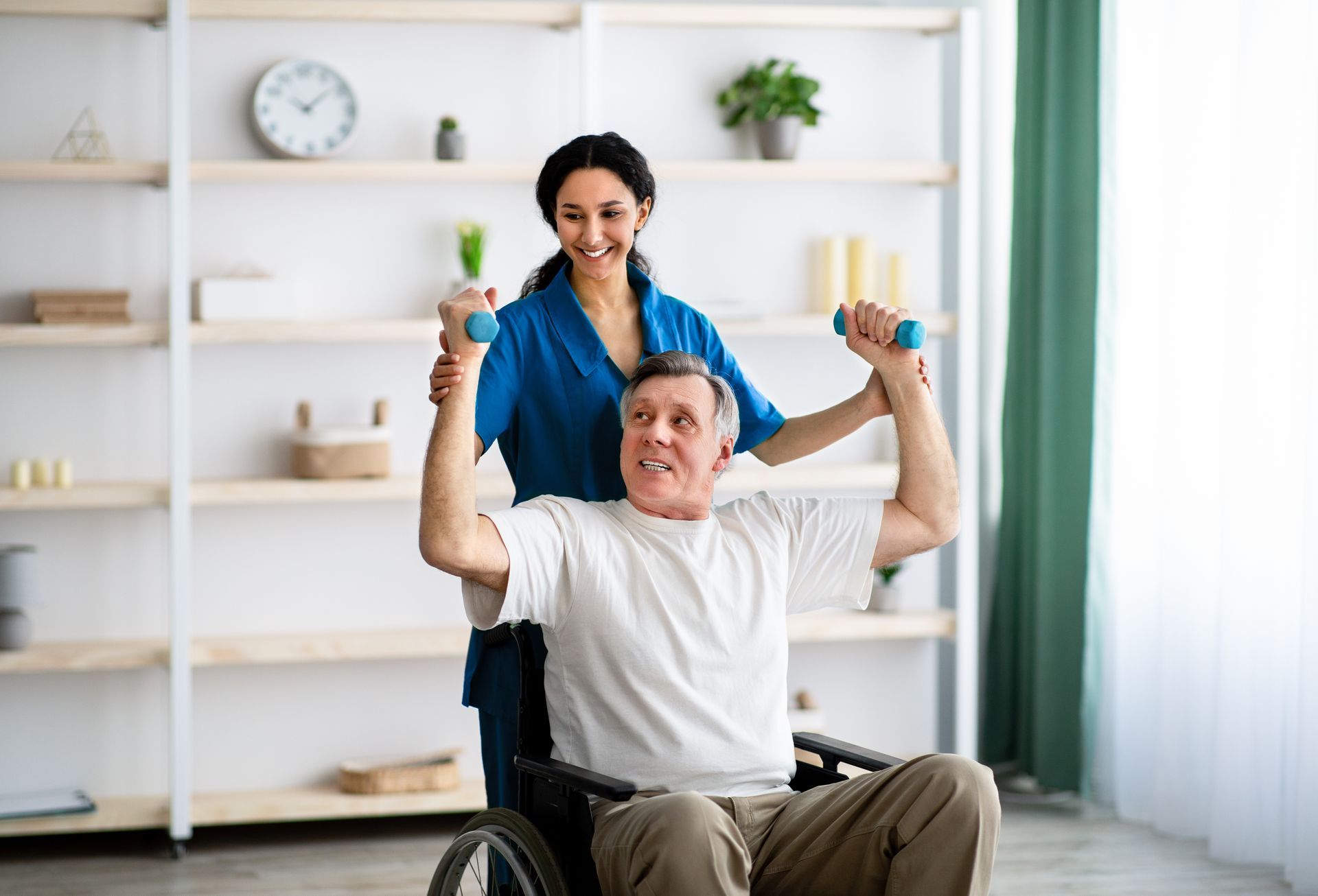 A nurse is helping an elderly man in a wheelchair lift dumbbells.