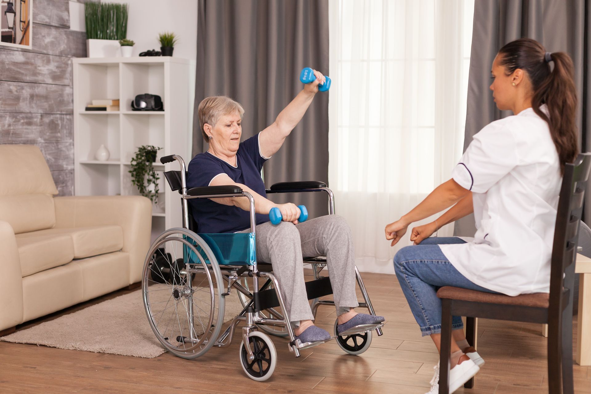 An elderly woman in a wheelchair is lifting dumbbells while a nurse watches.