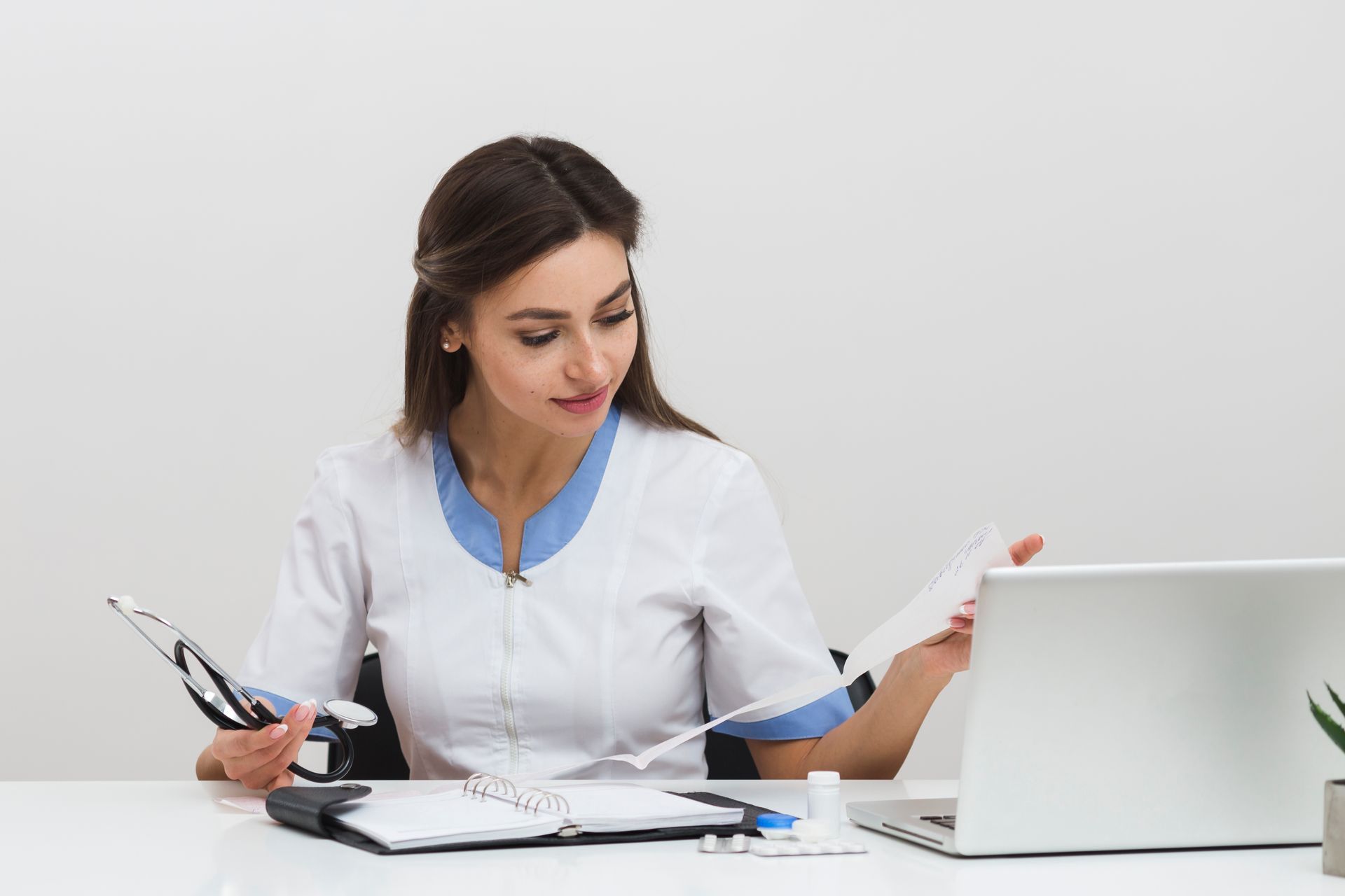 A female doctor is sitting at a desk with a laptop and a clipboard.