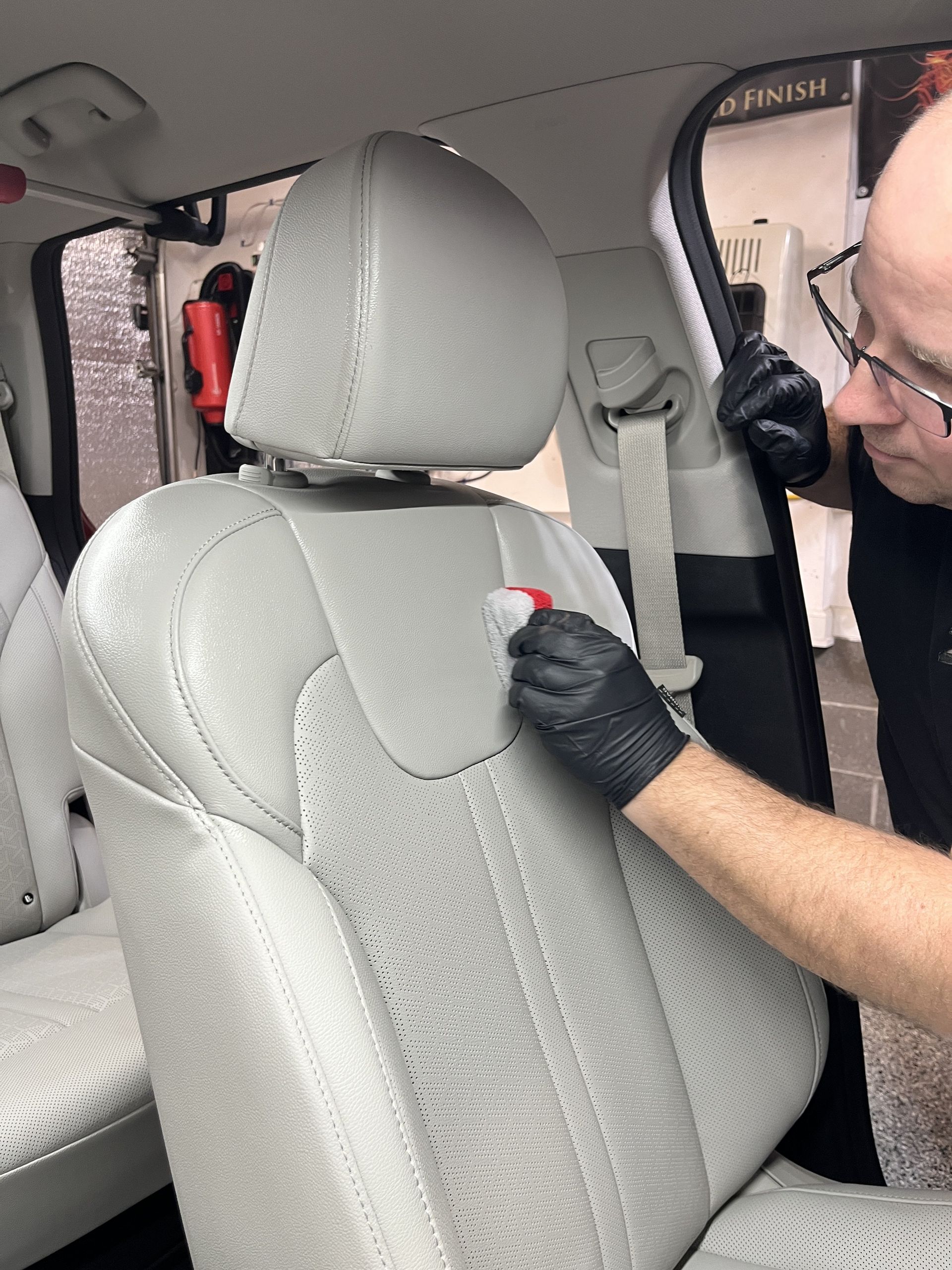 Person cleaning a light gray car seat with a brush, wearing black gloves, in a car interior.