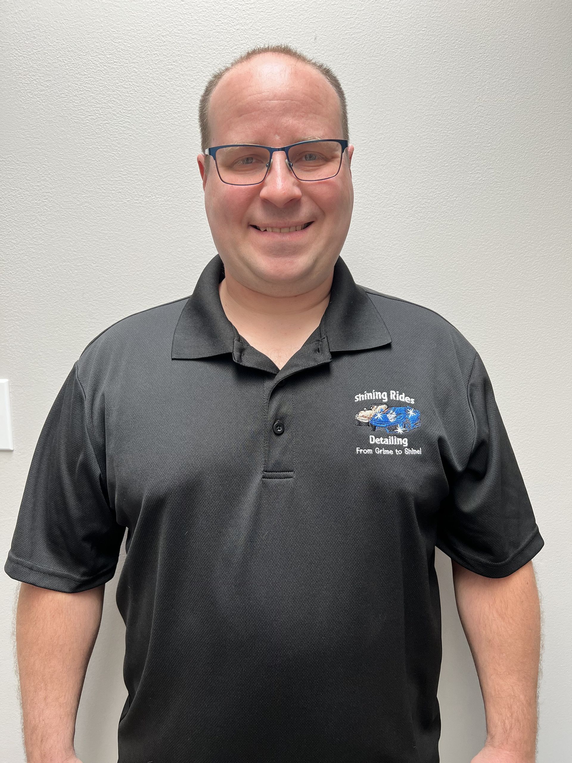 A man wearing glasses and a black polo shirt is smiling