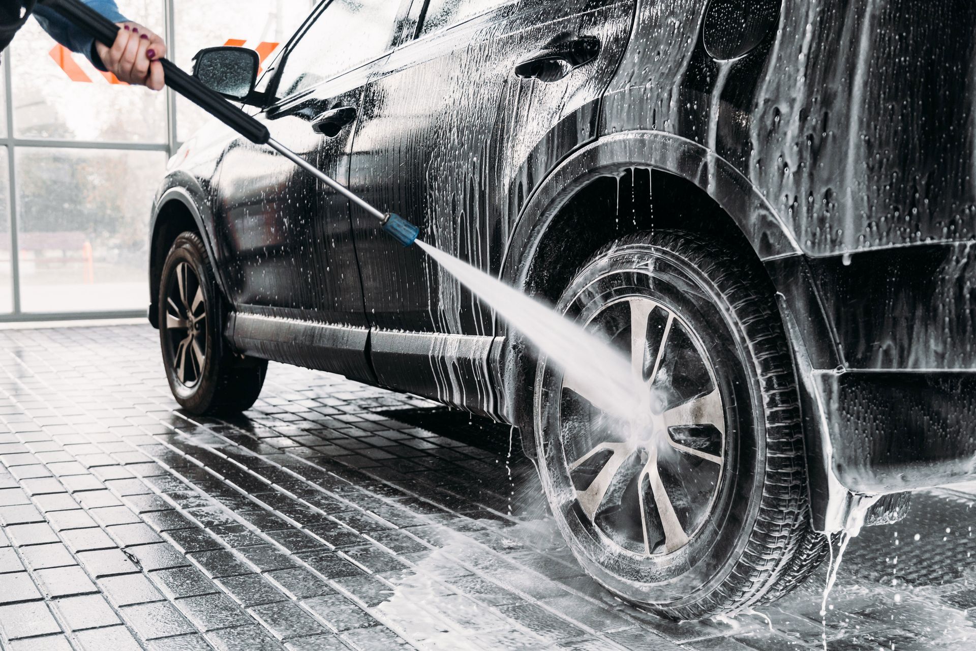 A person washing a black car with soapy water using a pressure washer at a car wash.