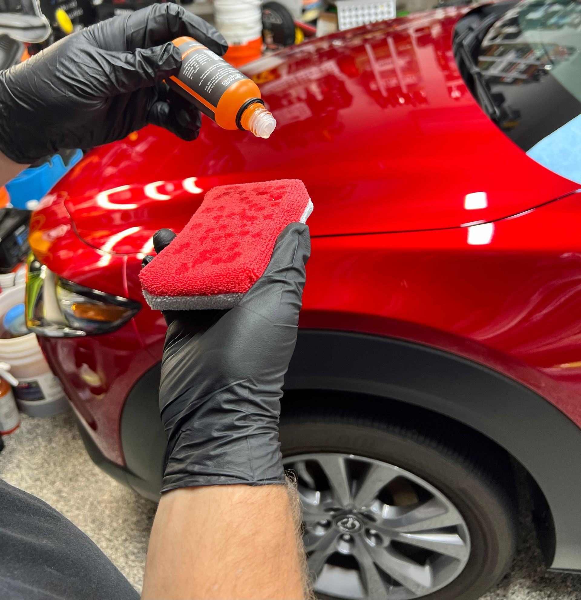 Person applying car wax to a red car with a sponge and bottle.