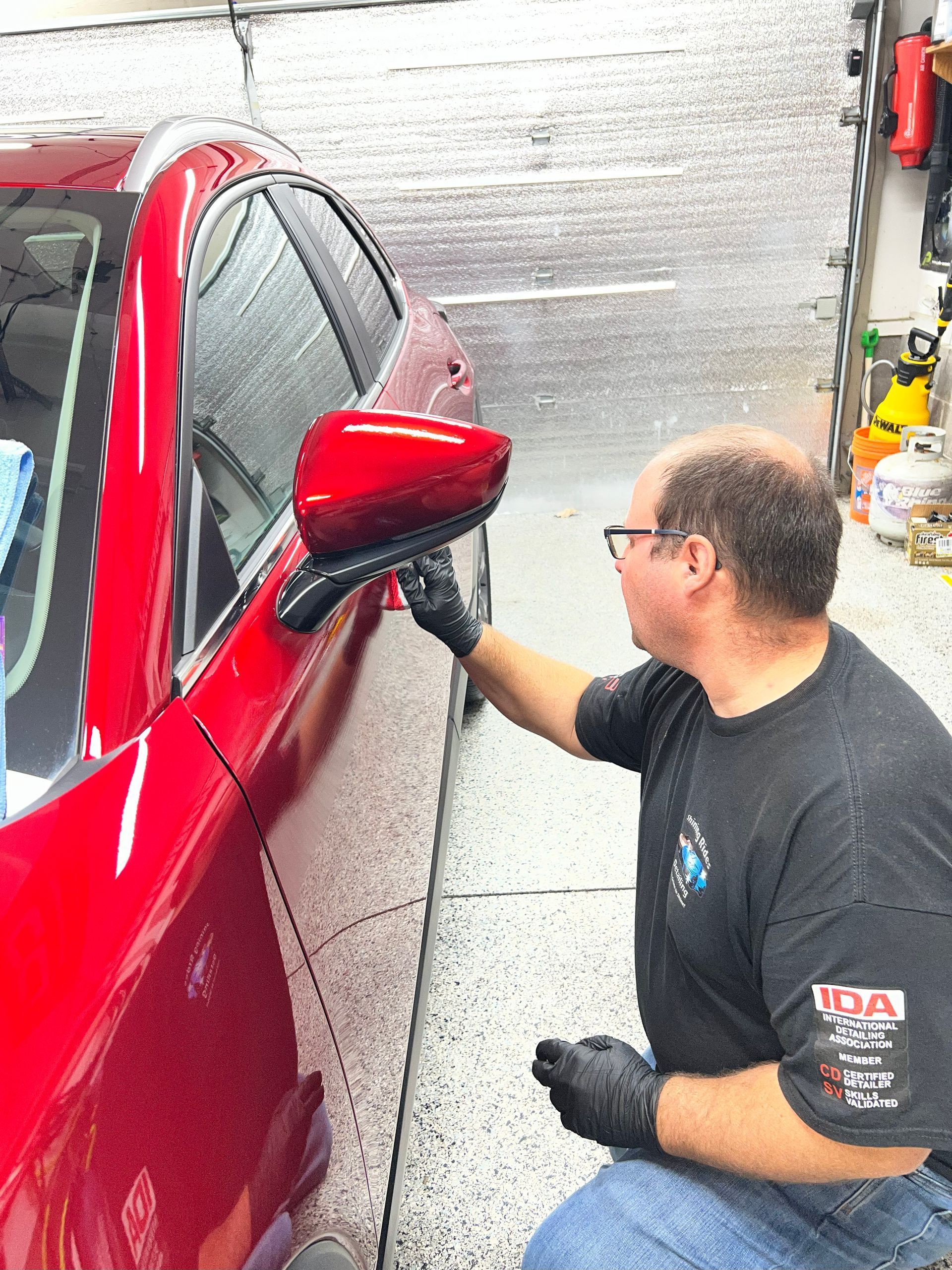 Person in black gloves wiping a red car mirror in a garage.