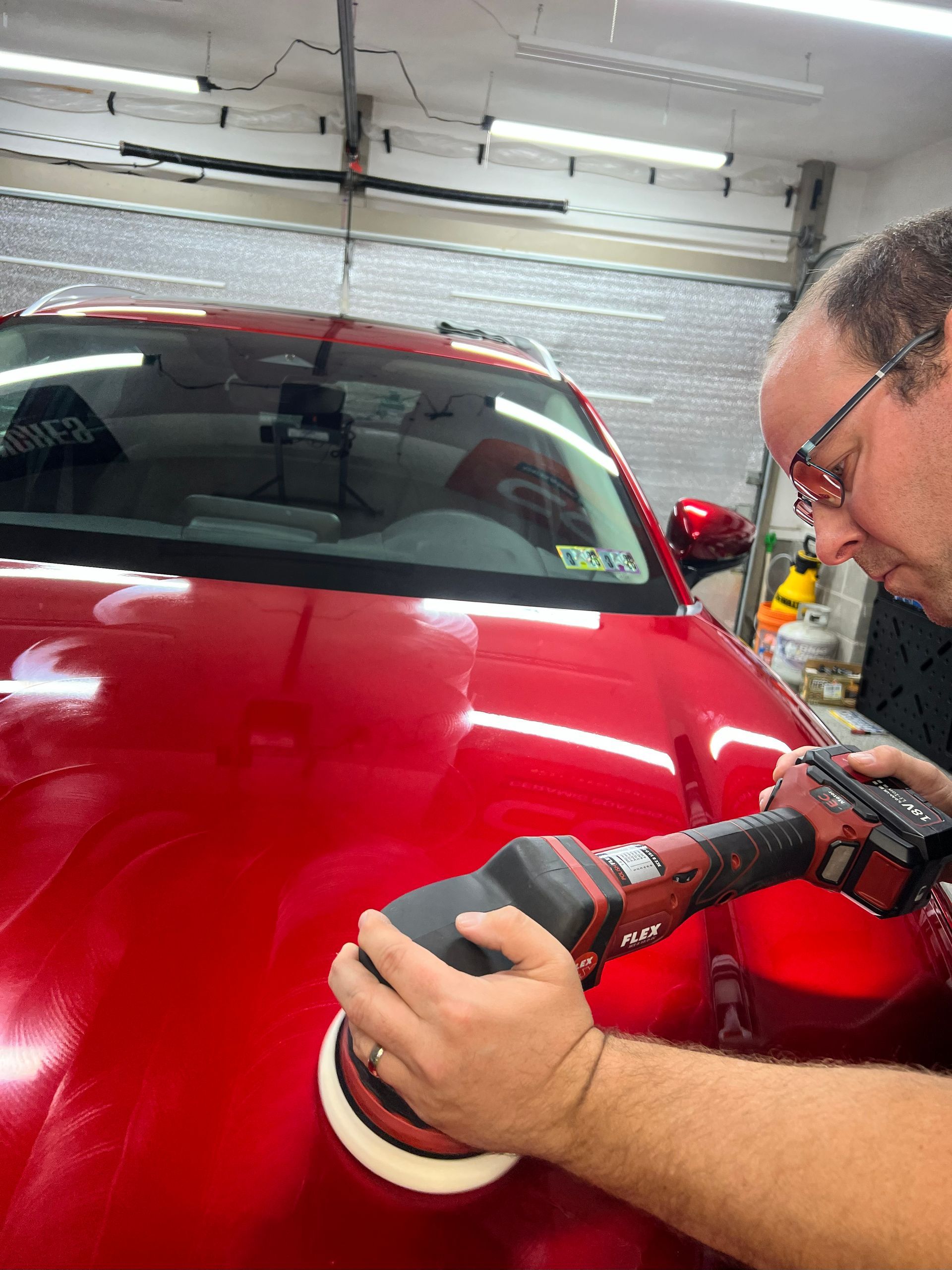 Man polishing a red car hood with a power buffer in a garage.