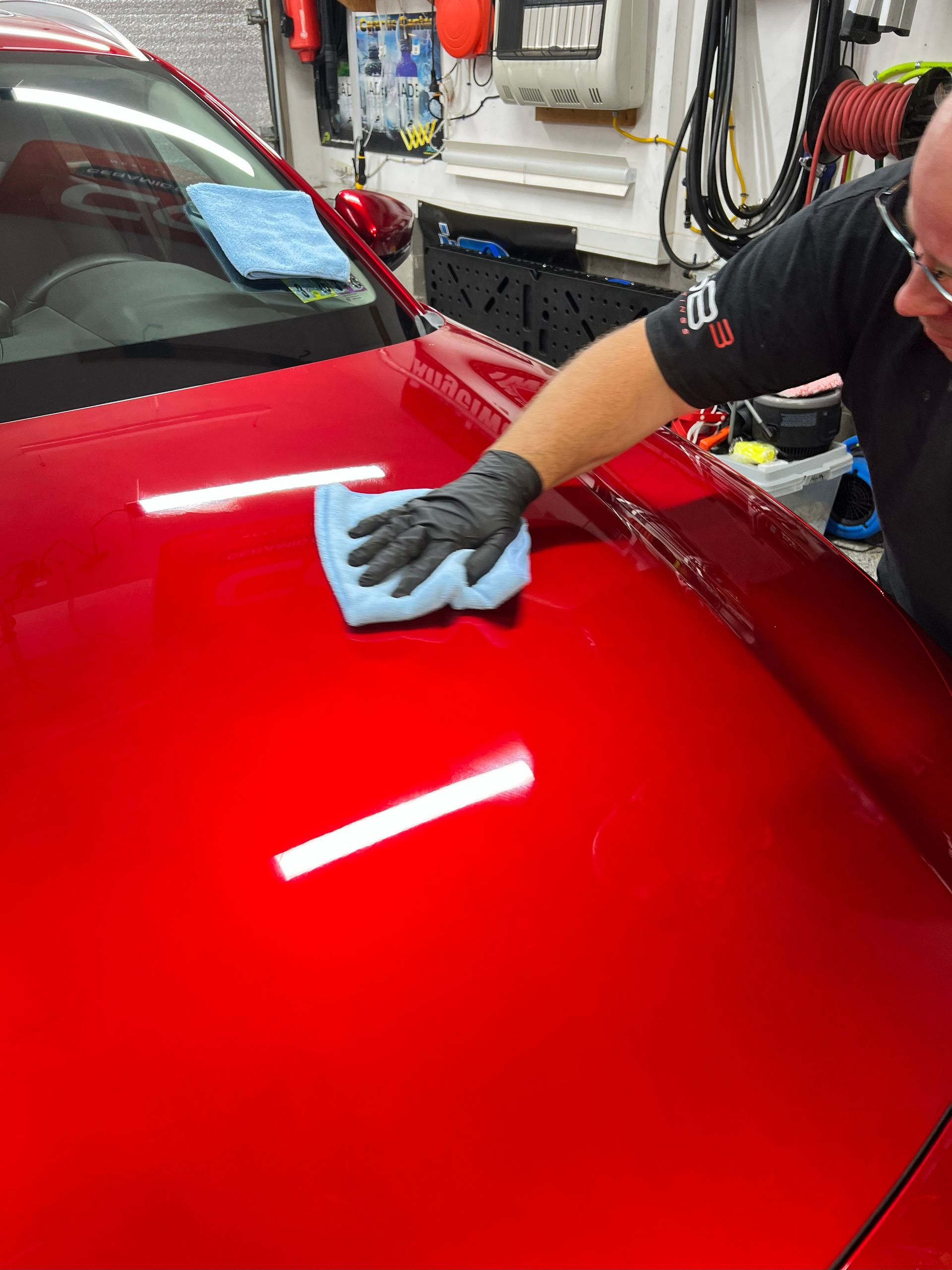 A person in gloves cleaning a shiny red car hood with a blue cloth.