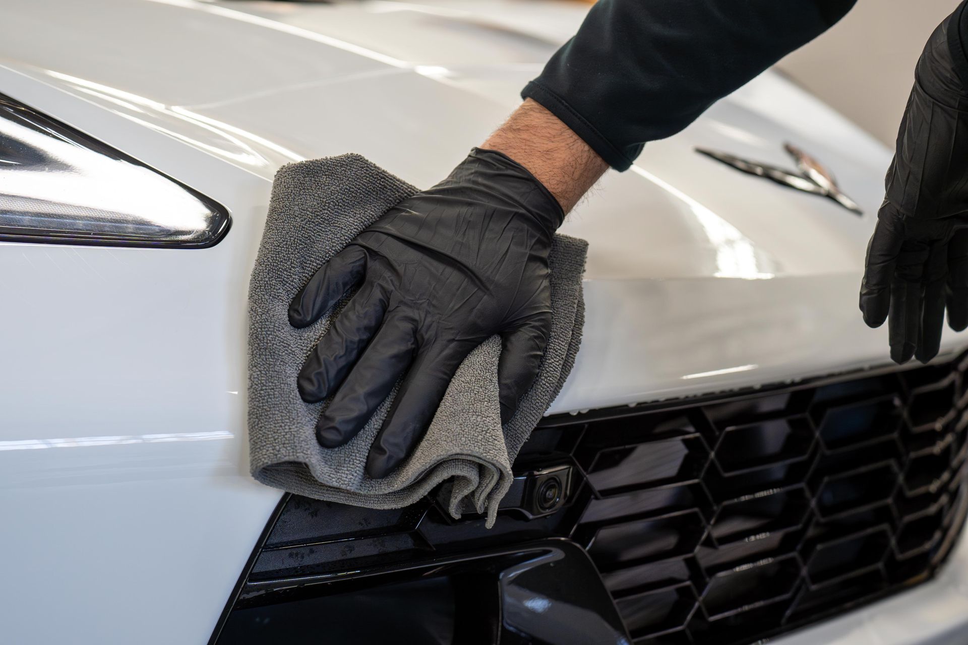 Person wearing black gloves wiping a white car's front with a gray microfiber cloth.